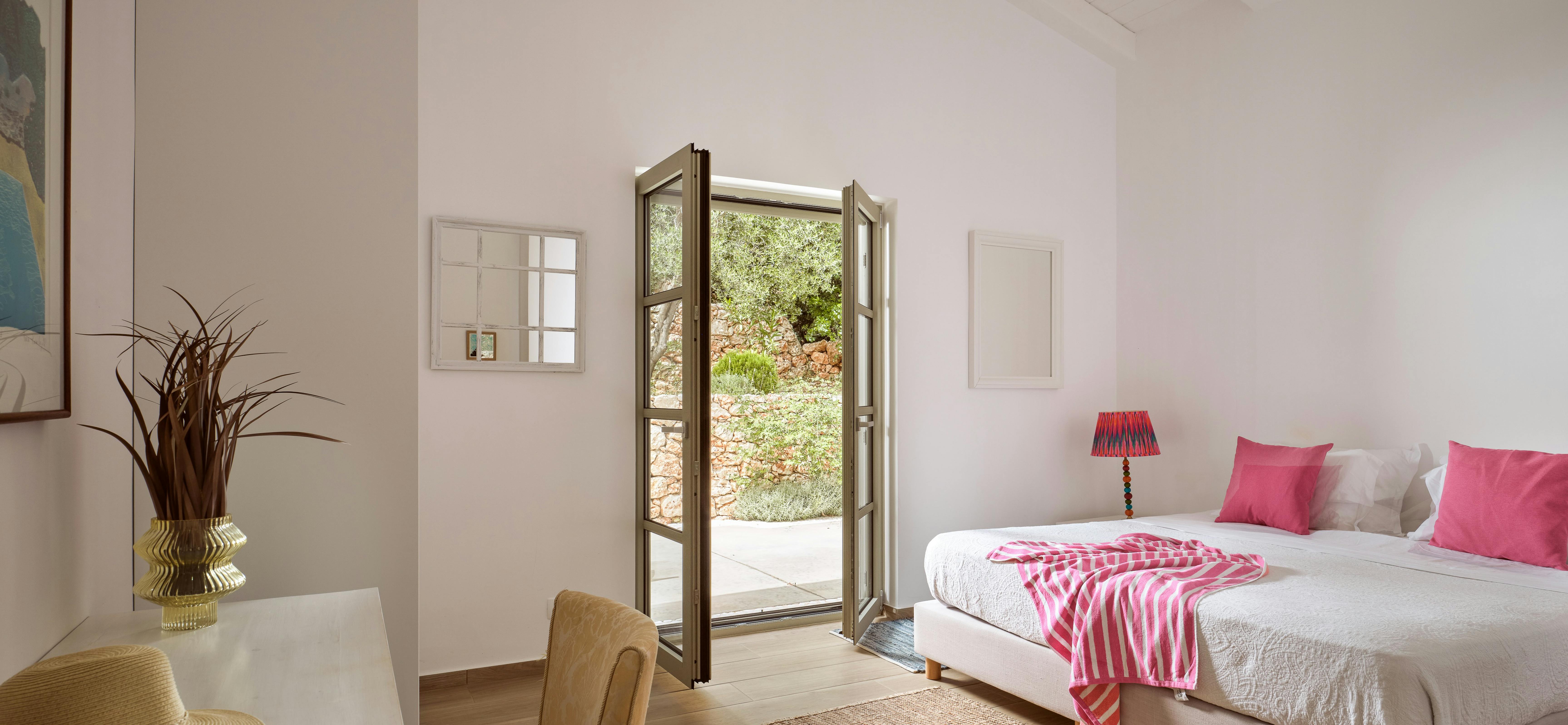 Bright bedroom with vaulted white ceiling opens directly to garden terrace through French doors. Pink accent pillows and striped throw add colour to the neutral palette and natural wood floors.