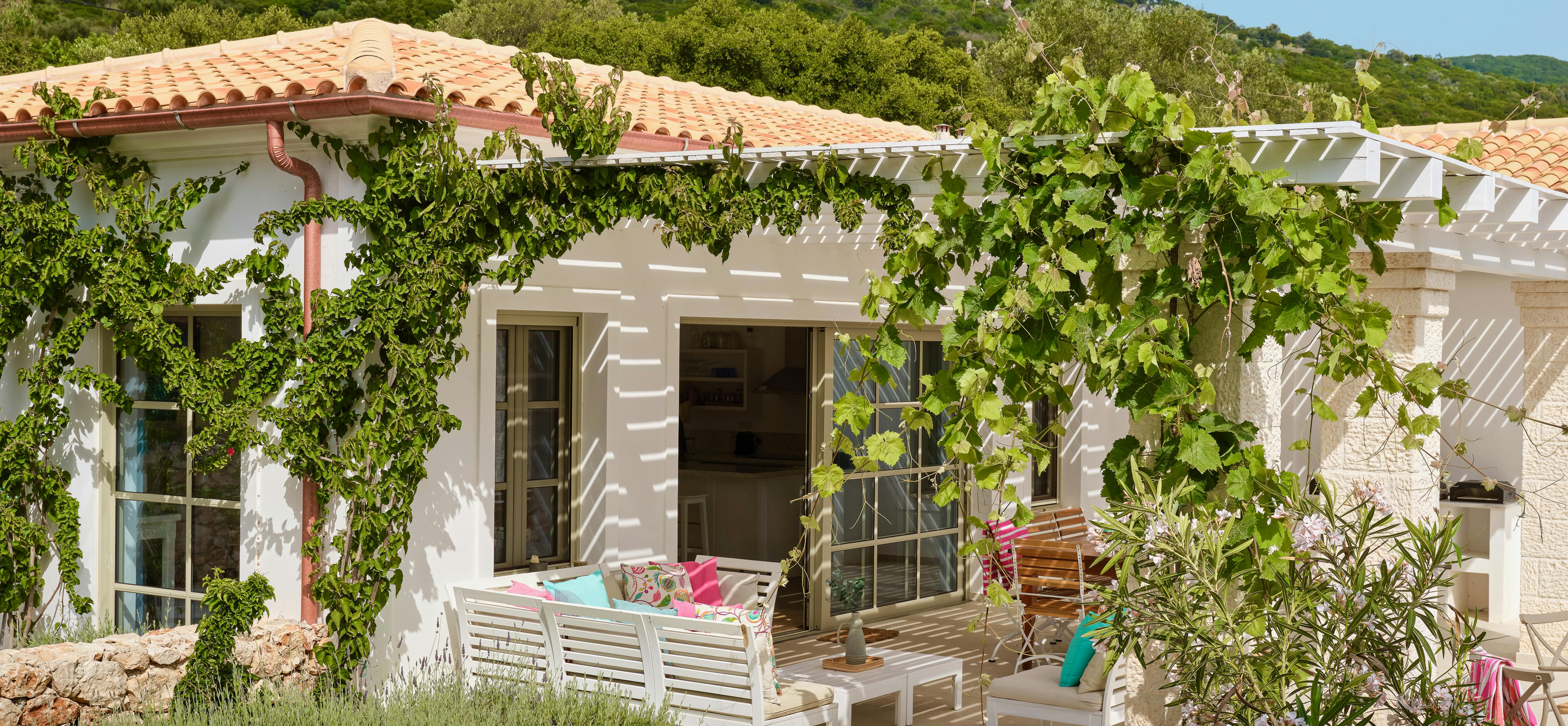 Traditional Mediterranean villa with terracotta roof tiles and vine-covered pergola terrace. White outdoor furniture and lush landscaping create inviting outdoor living space against rolling green hills.