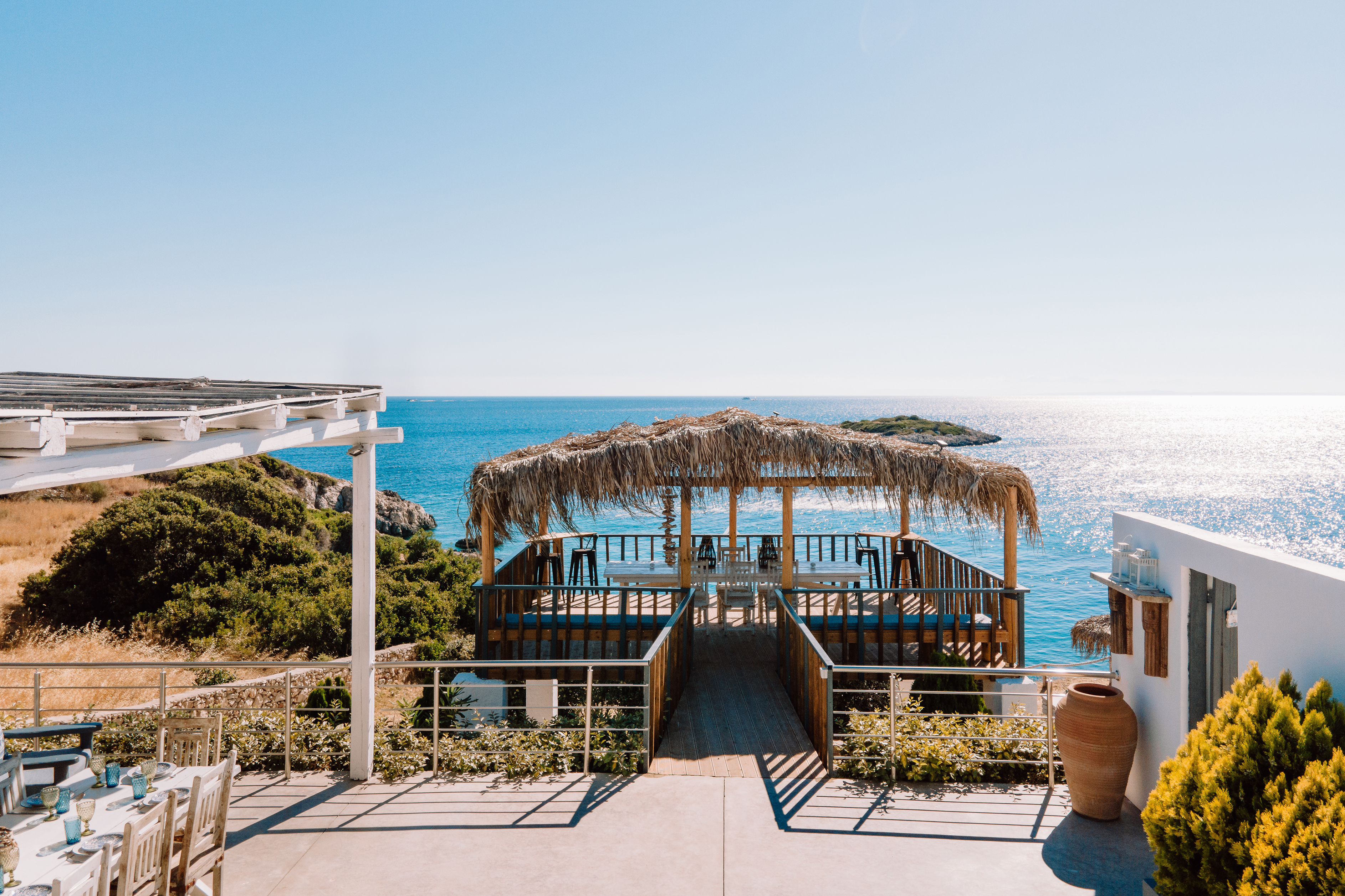 A seaside terrace features a traditional thatched-roof pavilion extending over crystal-clear turquoise waters. White dining furniture creates an elegant outdoor entertaining space.