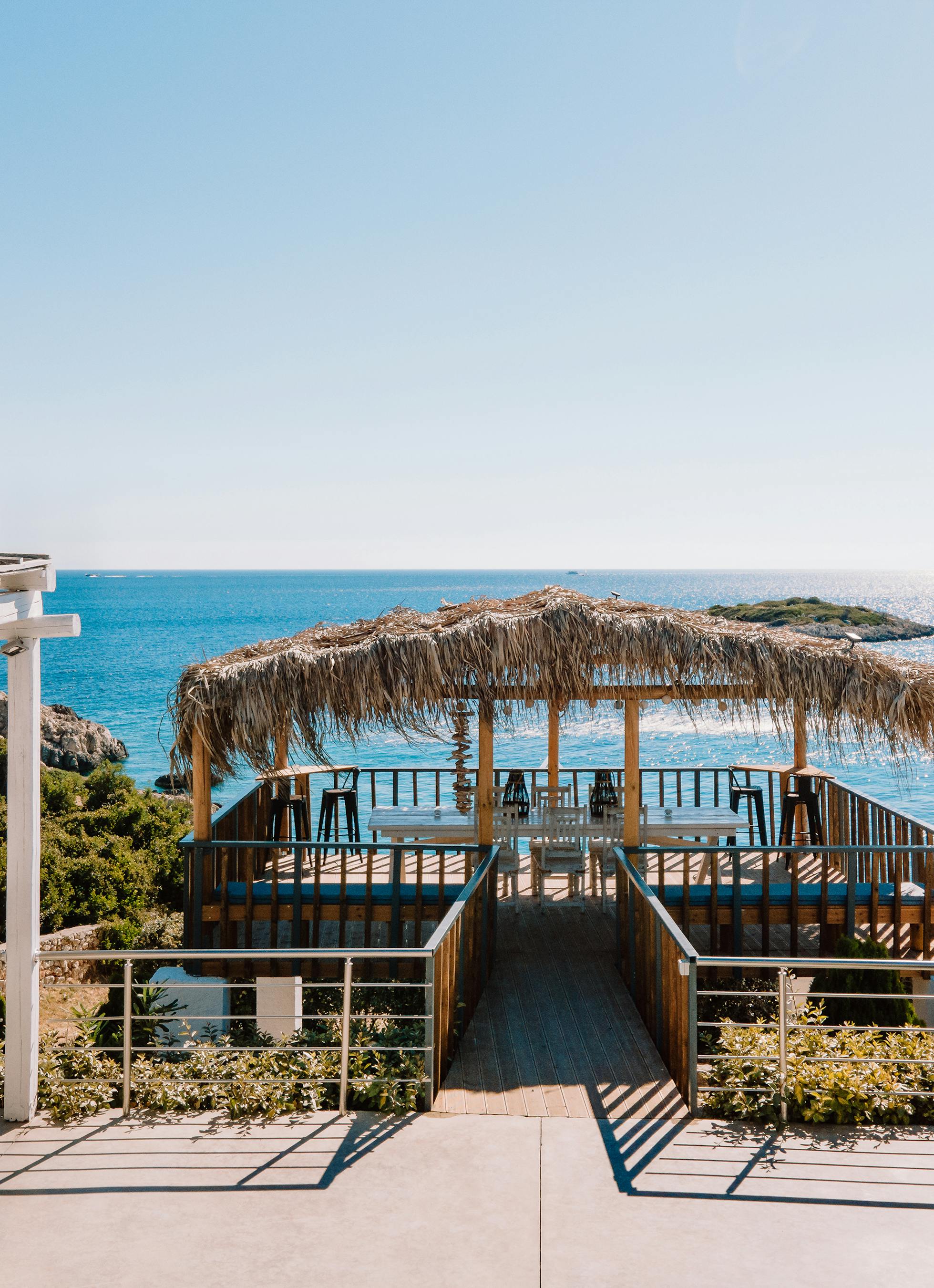 A seaside terrace features a traditional thatched-roof pavilion extending over crystal-clear turquoise waters. White dining furniture creates an elegant outdoor entertaining space.