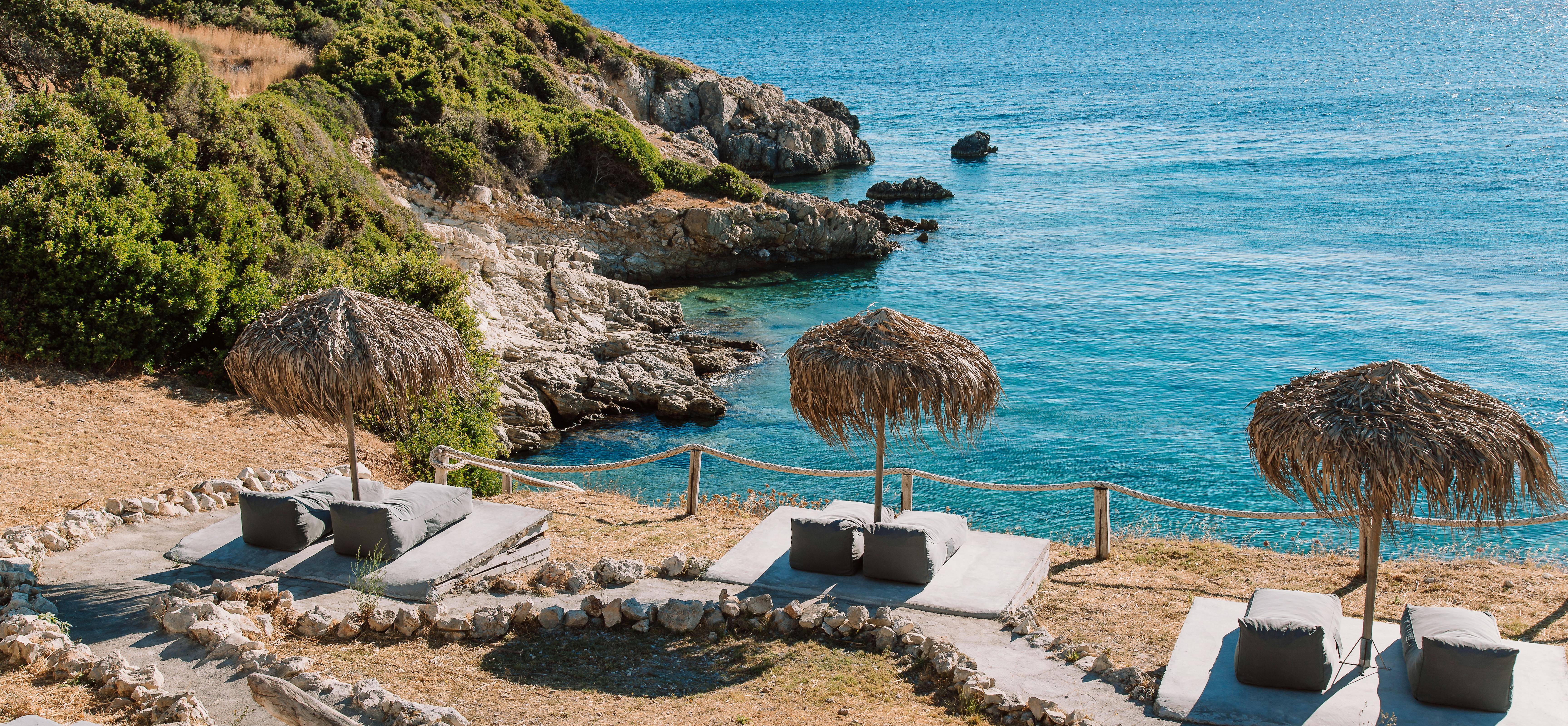 A clifftop terrace overlooks turquoise Mediterranean waters with built-in concrete seating platforms under traditional thatched umbrellas. The dramatic coastal setting features rocky outcrops and native vegetation along the shoreline.