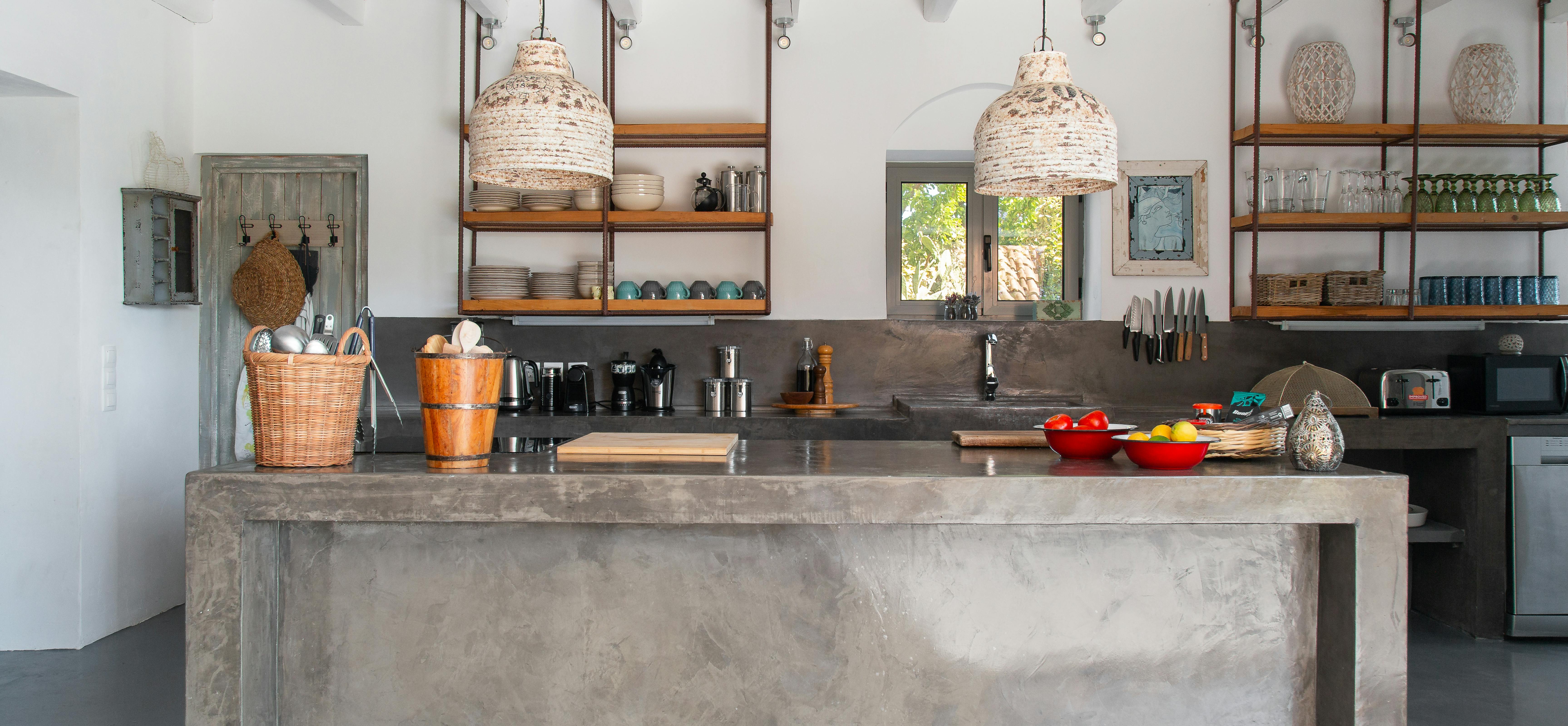 A modern kitchen features a concrete island with woven pendant lights and open wooden shelving displaying colourful ceramics. The minimalist design combines industrial materials with traditional Mediterranean elements and natural textures.