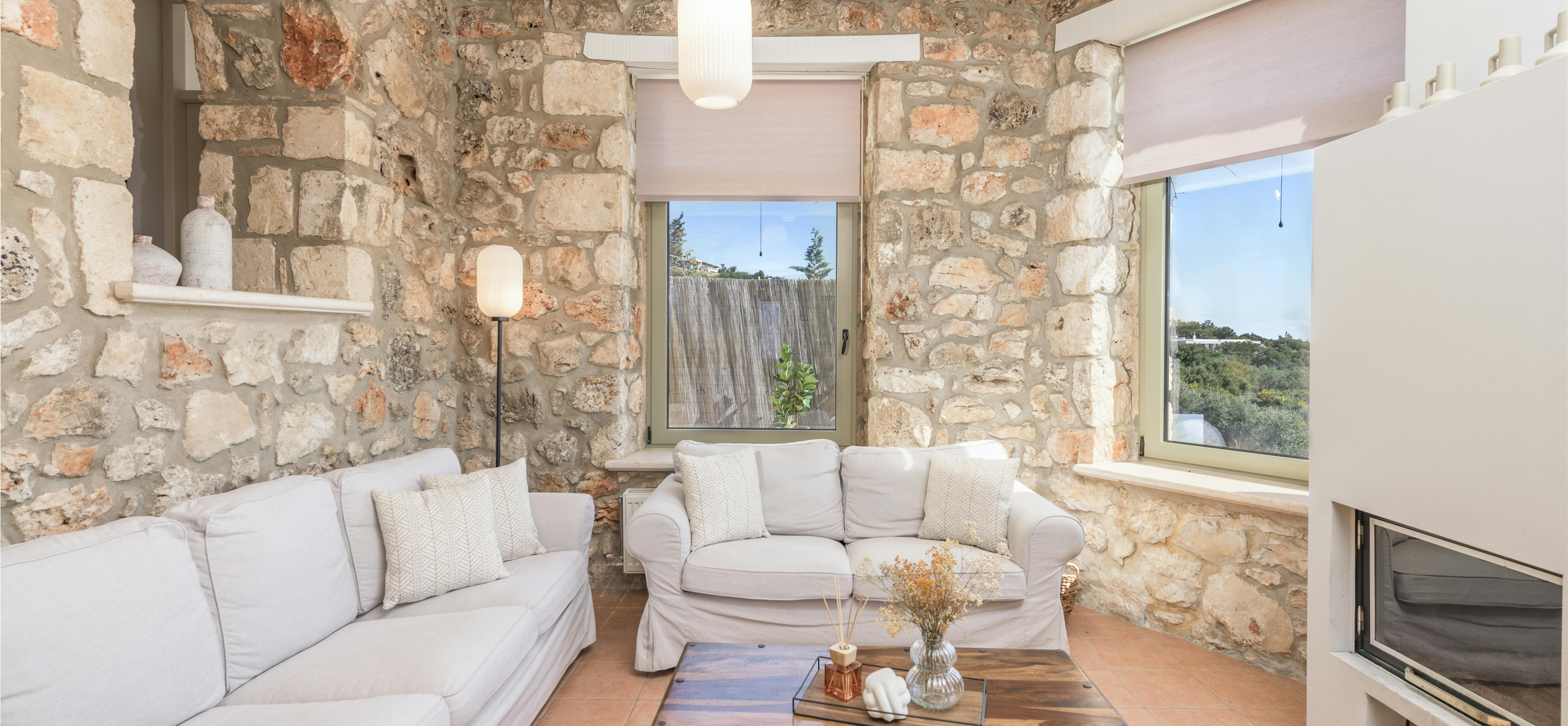 A living room features white slipcovered sofas arranged around a dark wooden coffee table beneath exposed ceiling beams. The neutral palette and natural textures create a relaxed atmosphere within the stone-walled space.