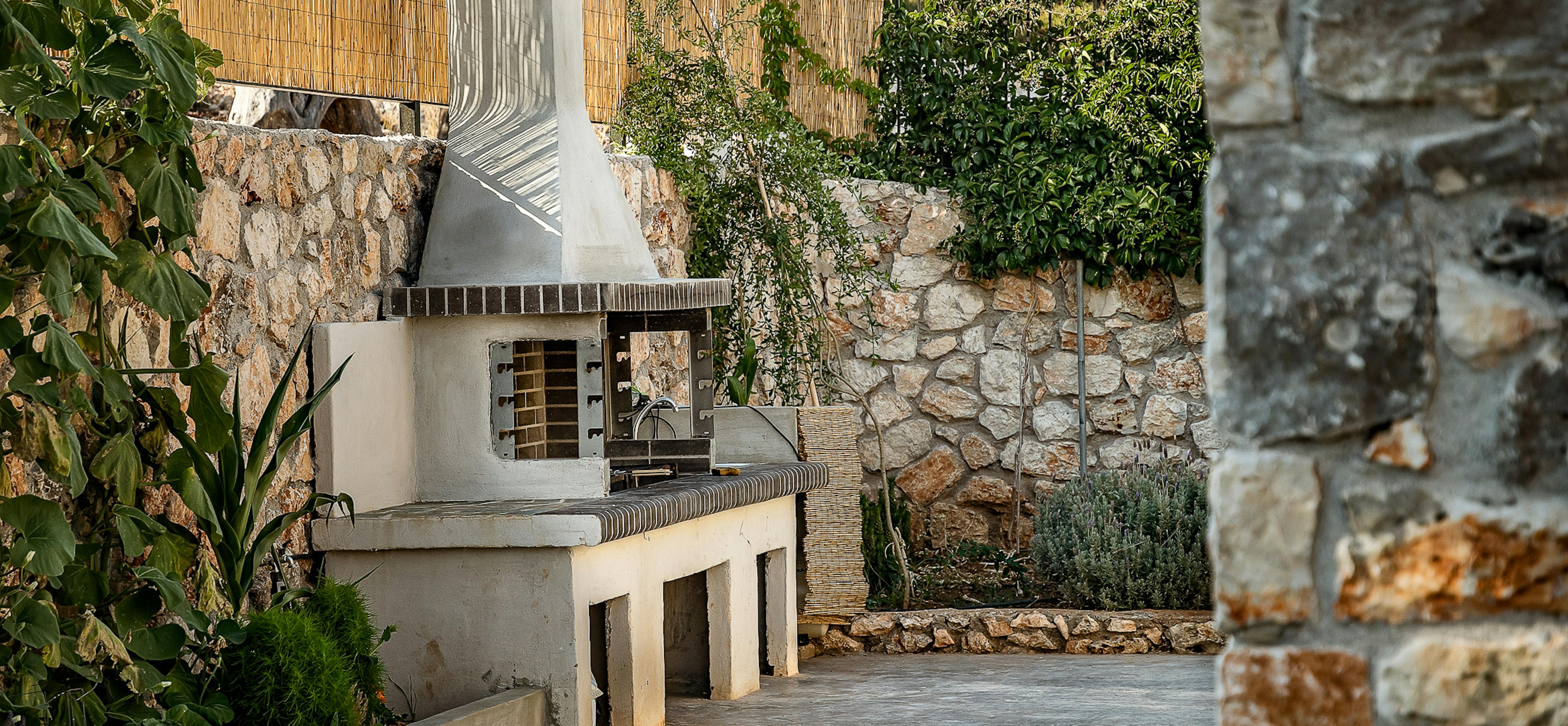 An outdoor kitchen area includes a built-in barbecue grill surrounded by stone walls and Mediterranean vegetation. The functional cooking space integrates seamlessly with the natural landscape and traditional stonework.