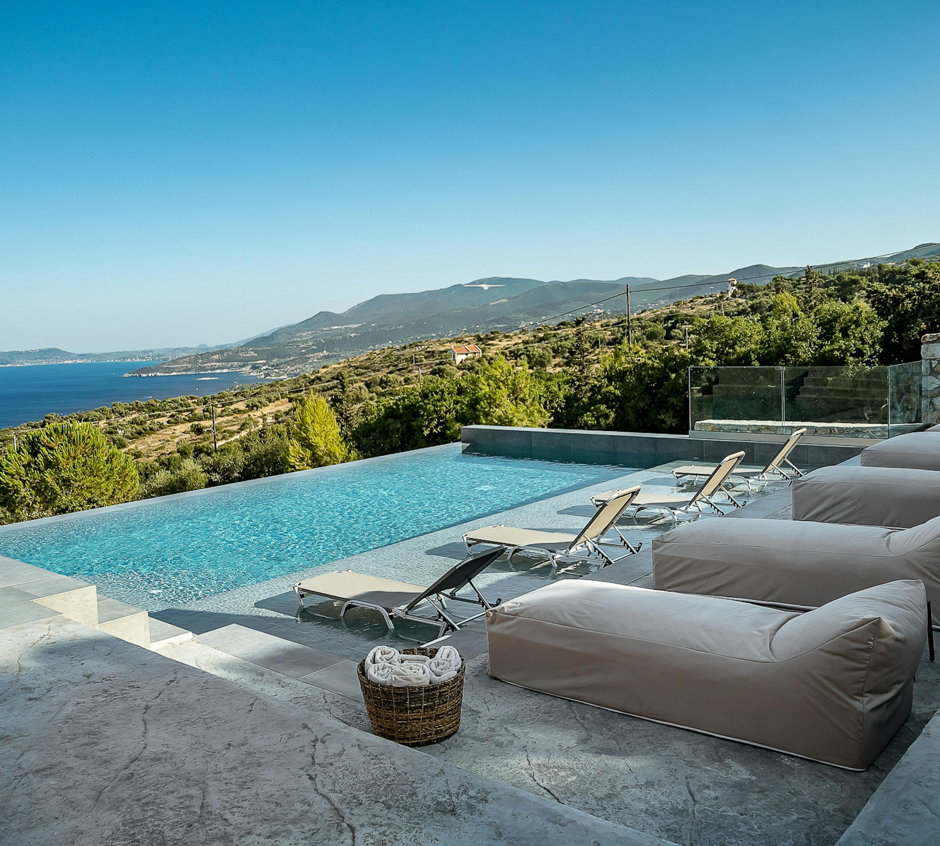 A hillside infinity pool terrace features large neutral cushions and lounge chairs positioned to maximise stunning Mediterranean sea views. The modern outdoor living space seamlessly blends luxury comfort with dramatic coastal scenery.