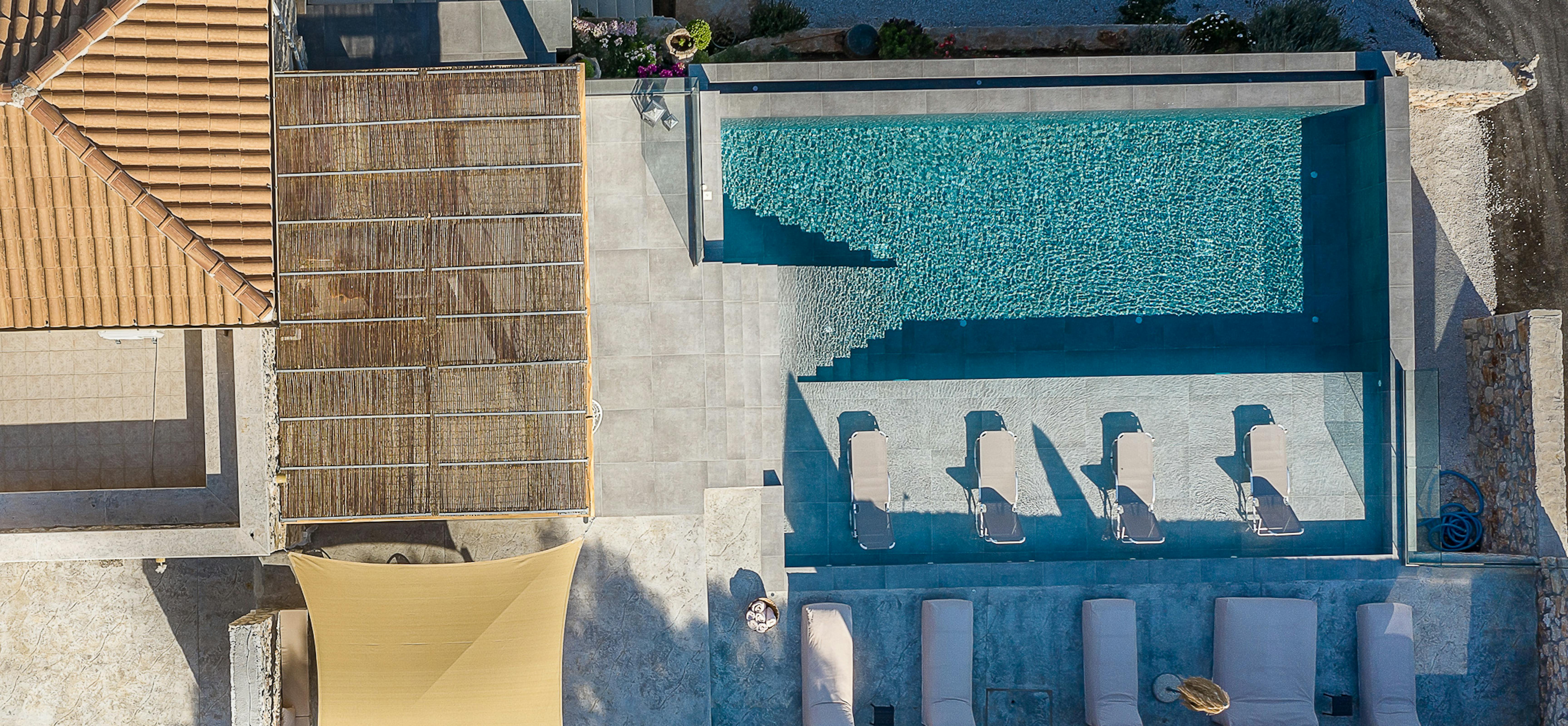 An aerial view shows a modern villa's multi-level pool area with white lounge chairs arranged on terraced decks. The infinity-edge swimming pool creates a striking geometric design against the hillside setting.