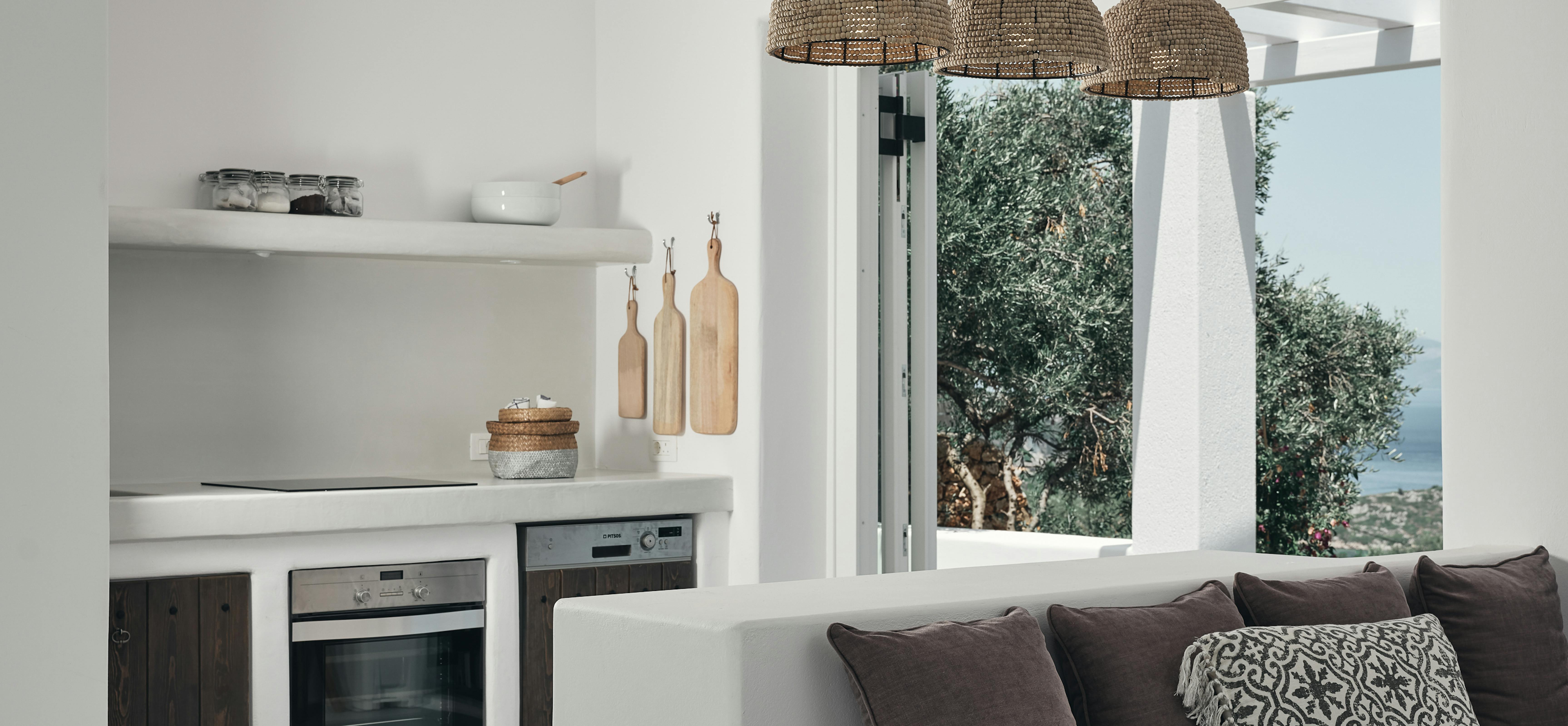 Open-plan white kitchen and living area with three woven pendant lights and built-in appliances. Grey sofa faces large windows overlooking olive trees and Mediterranean landscape.