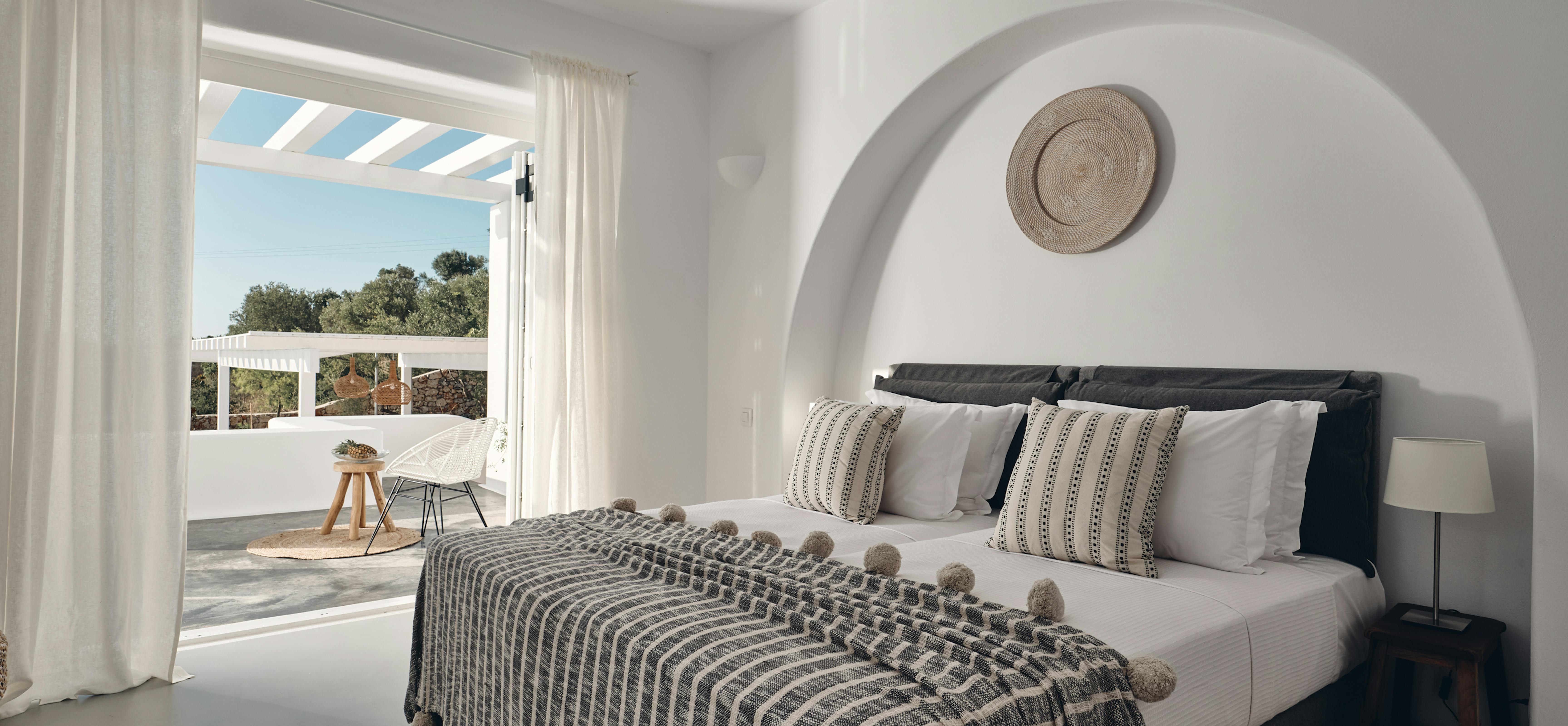 Serene bedroom with arched white alcove headboard and striped pom-pom throw opens to private terrace through floor-to-ceiling curtains. Woven basket artwork and neutral textiles create calming Mediterranean retreat atmosphere.