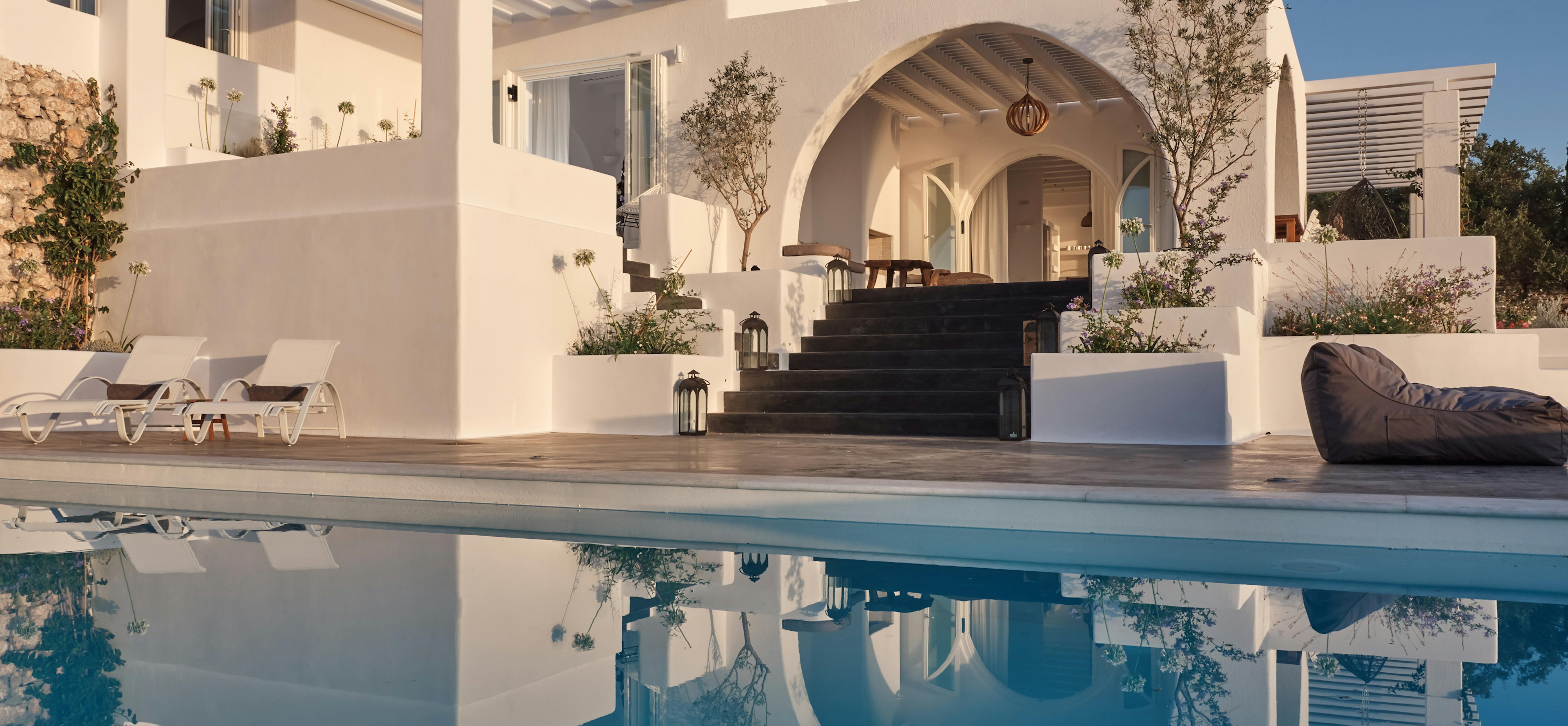 Pristine infinity pool perfectly reflects white villa architecture with dramatic arched entrance and stone staircase. Multi-level terraces and pergola structures create stunning geometric composition against clear blue sky.