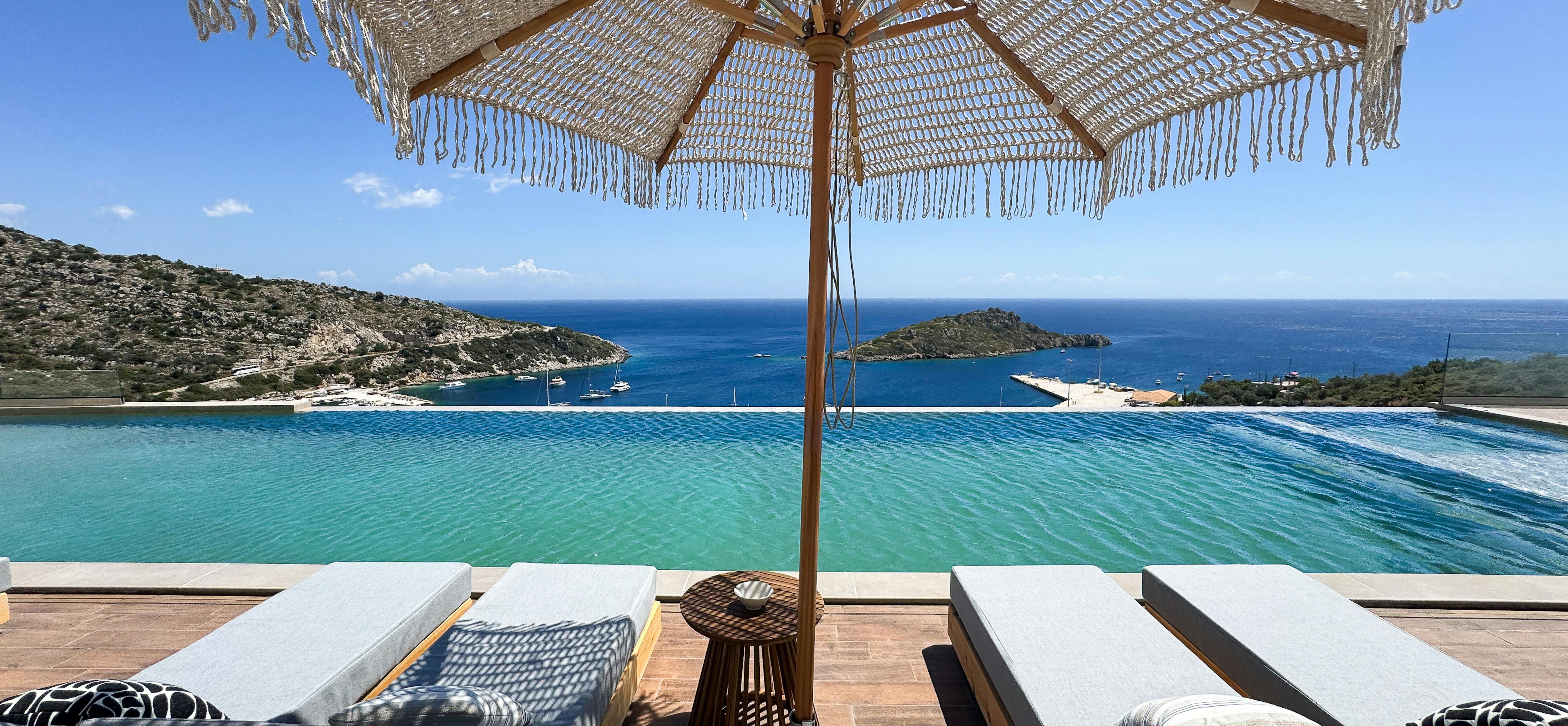 Luxury poolside lounging area with woven umbrella and teak furniture frames stunning sea views toward distant coastal islands. Grey cushioned sun beds and wicker side table create intimate outdoor relaxation space.