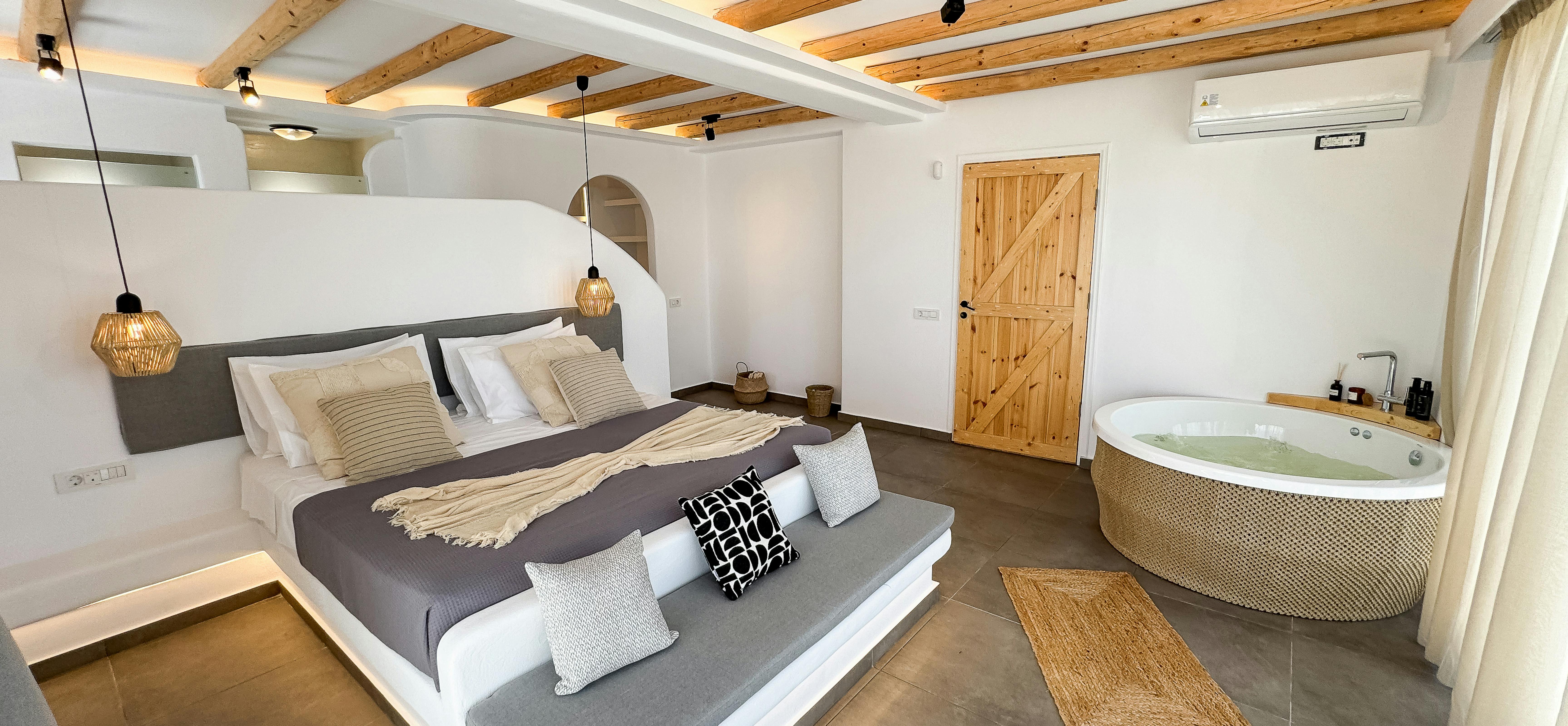 Luxury master suite with platform bed, exposed ceiling beams, and corner jacuzzi tub overlooks private terrace. Woven pendant lights and natural textiles enhance the resort-like bedroom atmosphere.