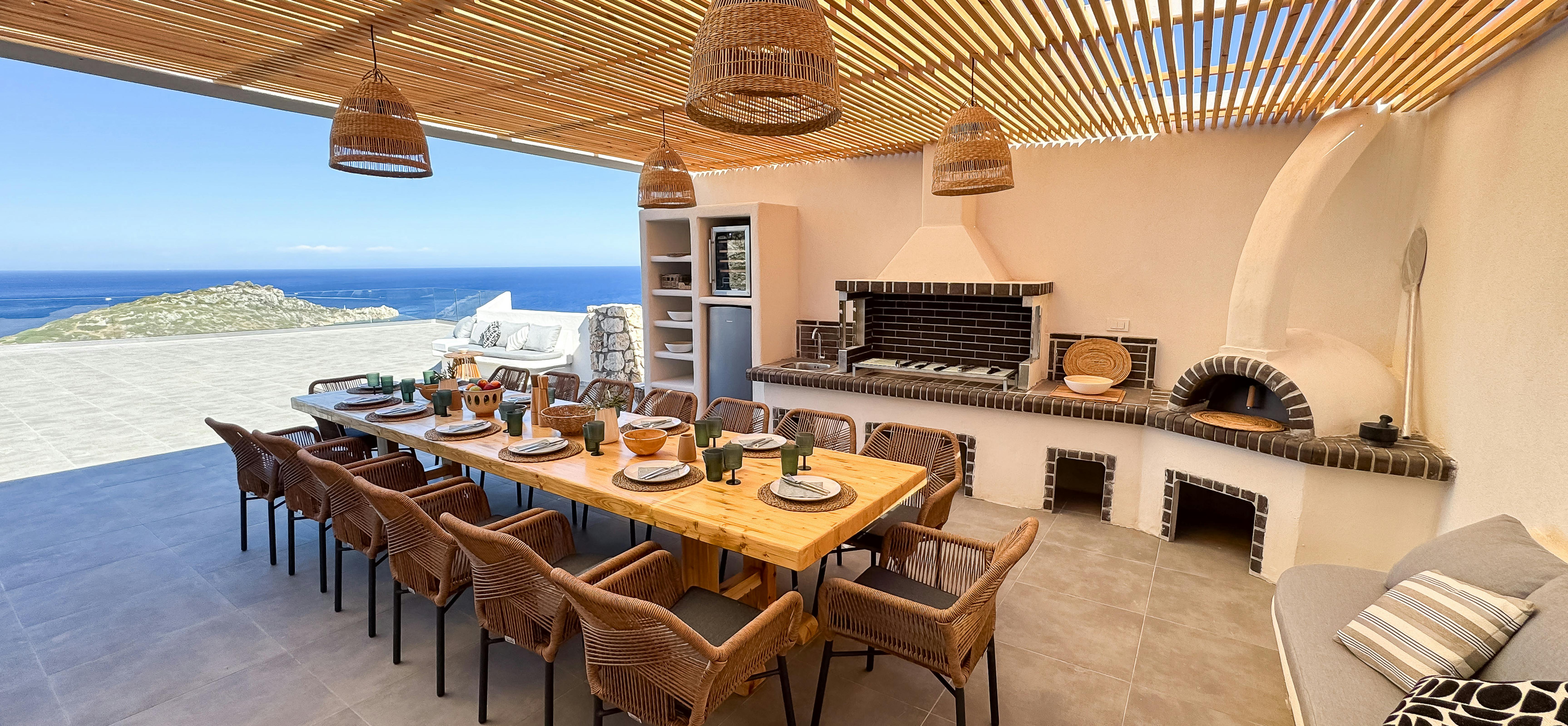 Covered outdoor dining pavilion with wood-fired oven, built-in grills, and woven pendant lights overlooks infinity pool and sea. Natural stone construction and teak furniture create perfect alfresco entertaining space.