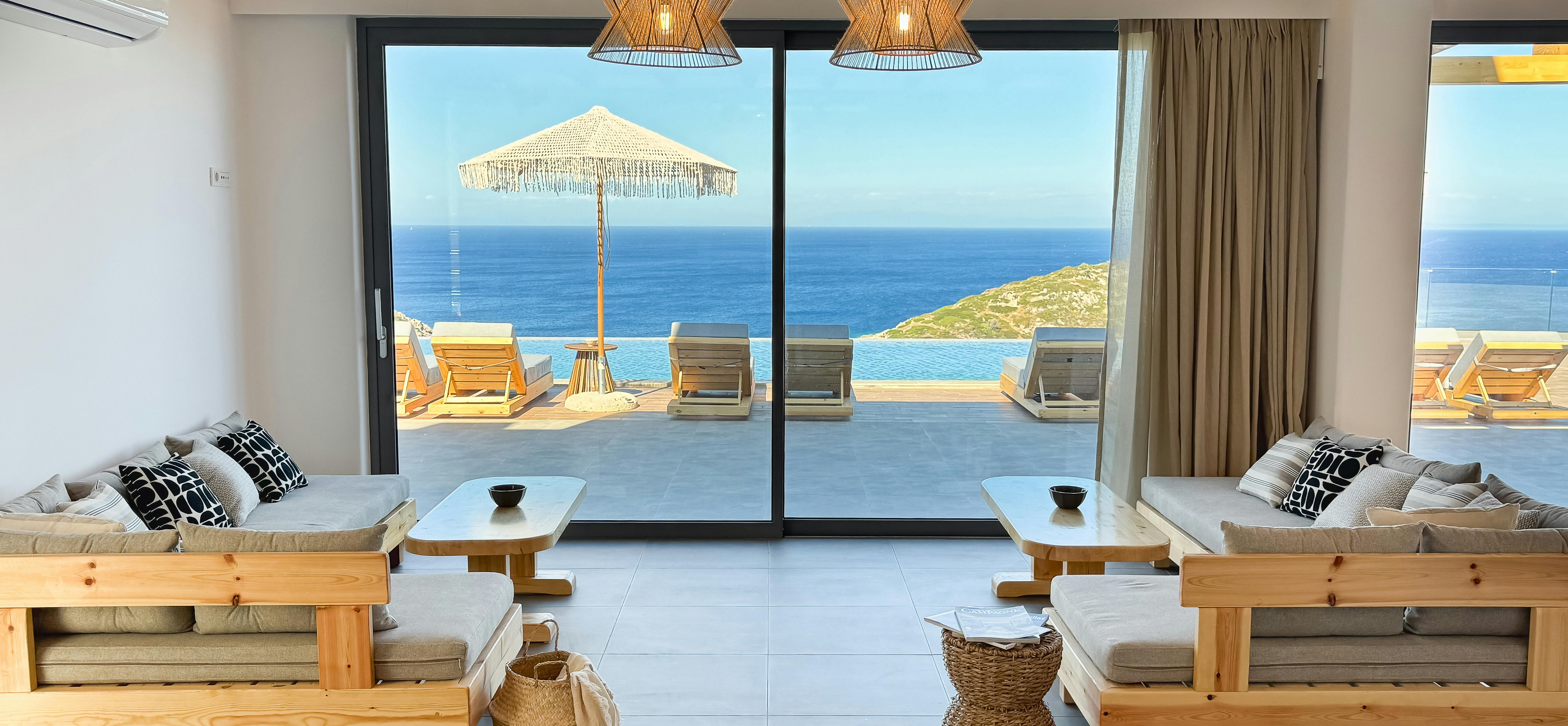 Living room with natural wood sectional seating, exposed beams, and woven pendant lights opens completely to infinity pool terrace. Floor-to-ceiling windows maximise Mediterranean sea views from every angle.