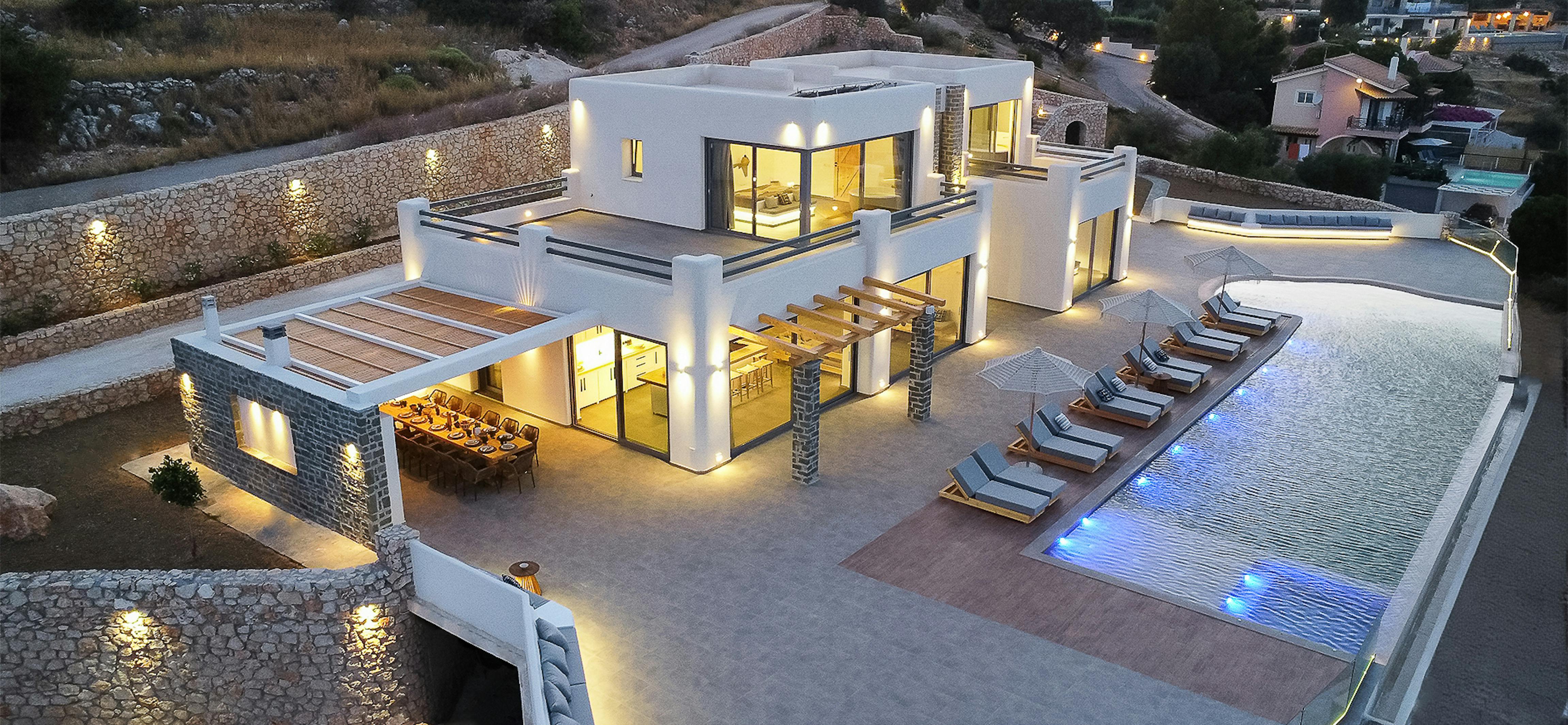 Aerial evening view reveals complete luxury villa complex with illuminated pool, multi-level terraces, and stone retaining walls integrated into Mediterranean hillside. Architectural lighting highlights the geometric white structures against landscape.