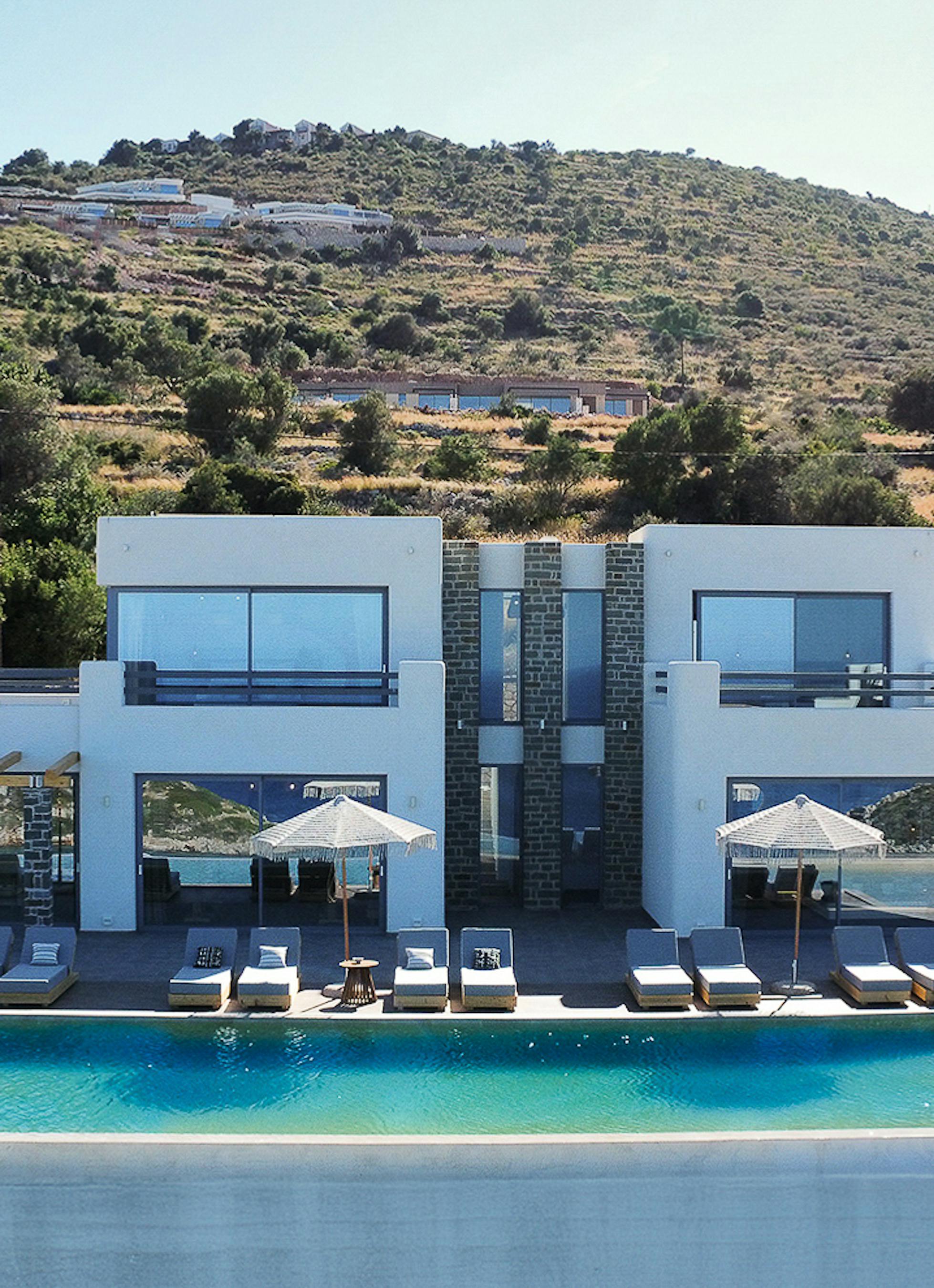 Stunning aerial view of complete villa complex showing curved infinity pool, multiple terraces, and integration with natural hillside landscape. Modern white architecture contrasts beautifully with surrounding Mediterranean countryside.