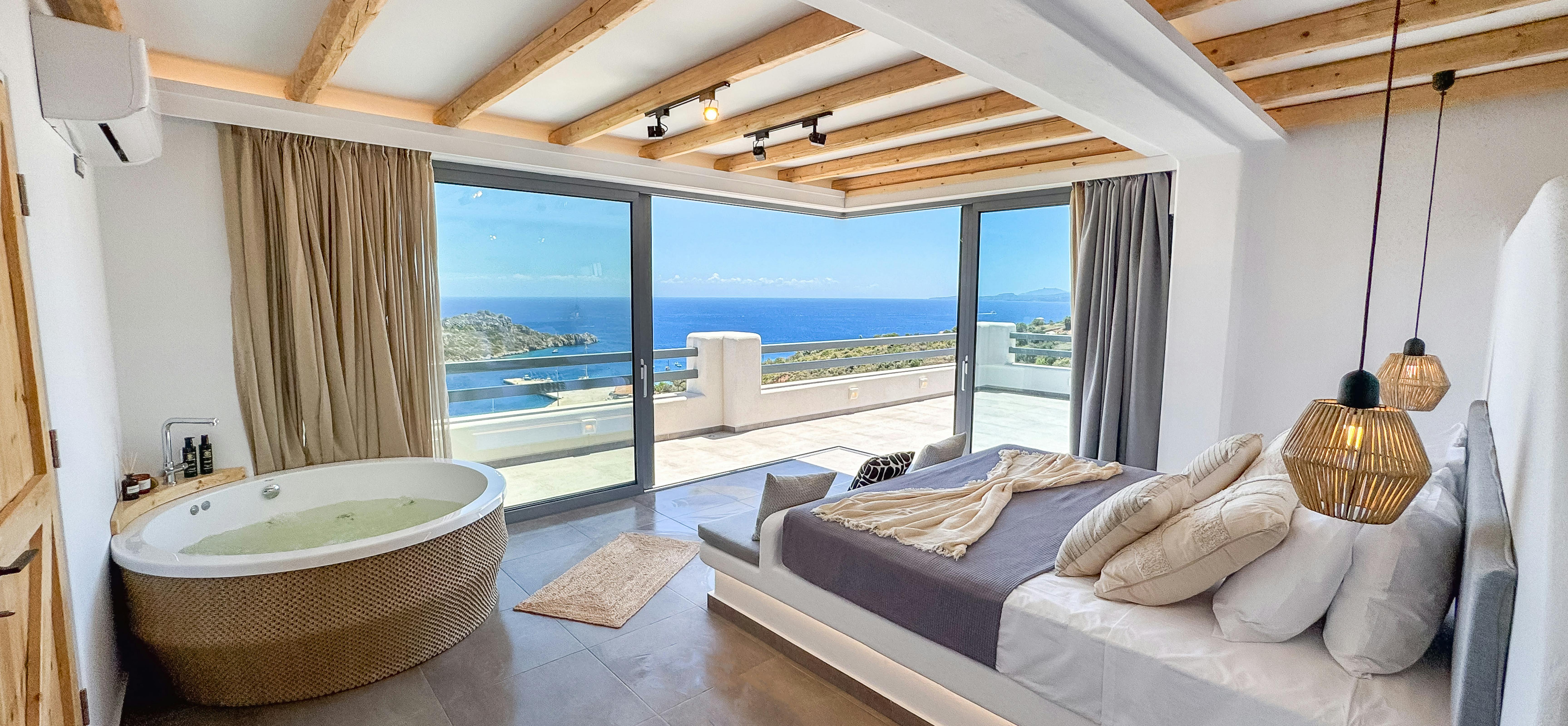 Luxury master suite with corner jacuzzi tub, exposed wood beam ceiling, and panoramic sea views through floor-to-ceiling windows. Platform bed and woven pendant lights create spa-like atmosphere with direct terrace access.