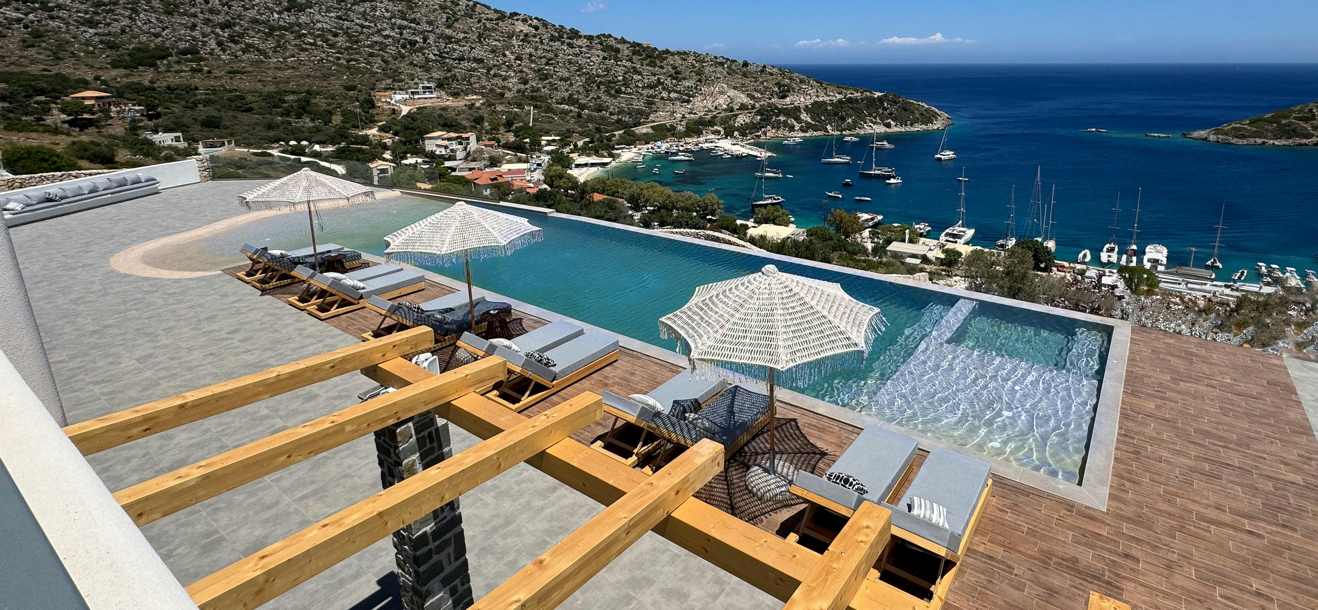 Dramatic clifftop pool terrace with infinity edge overlooking natural harbour filled with anchored sailboats and yachts. Wooden pergola structure frames lounging areas with woven umbrellas providing shade above the stunning coastal panorama.