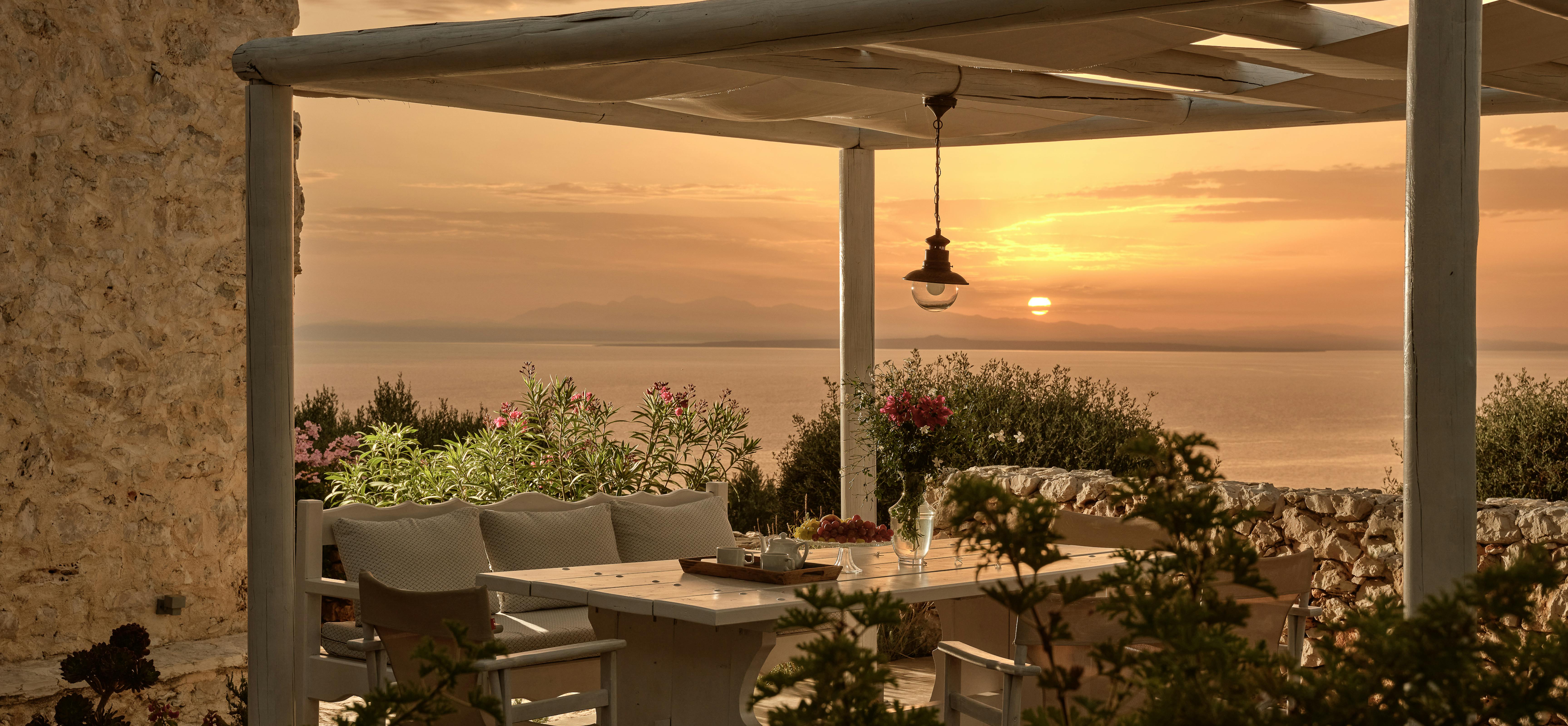 Romantic sunset terrace dining with white pergola overlooking golden Mediterranean sea horizon. Stone walls and flowering plants frame intimate table setting under pendant light as dramatic clouds reflect warm evening colours.
