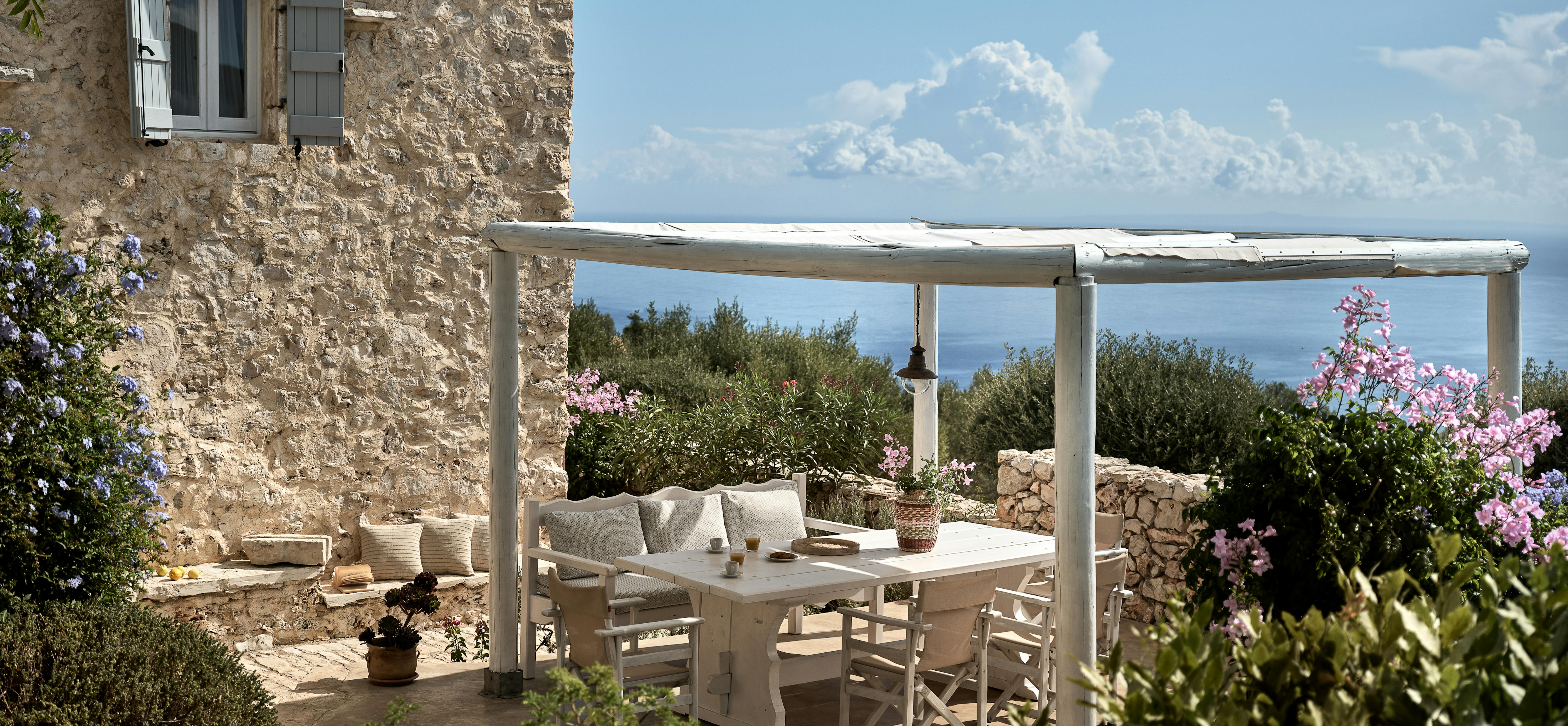 Traditional stone farmhouse terrace with white pergola and outdoor seating surrounded by blooming purple flowers. Grey shuttered windows and natural stone walls create authentic Mediterranean countryside charm.