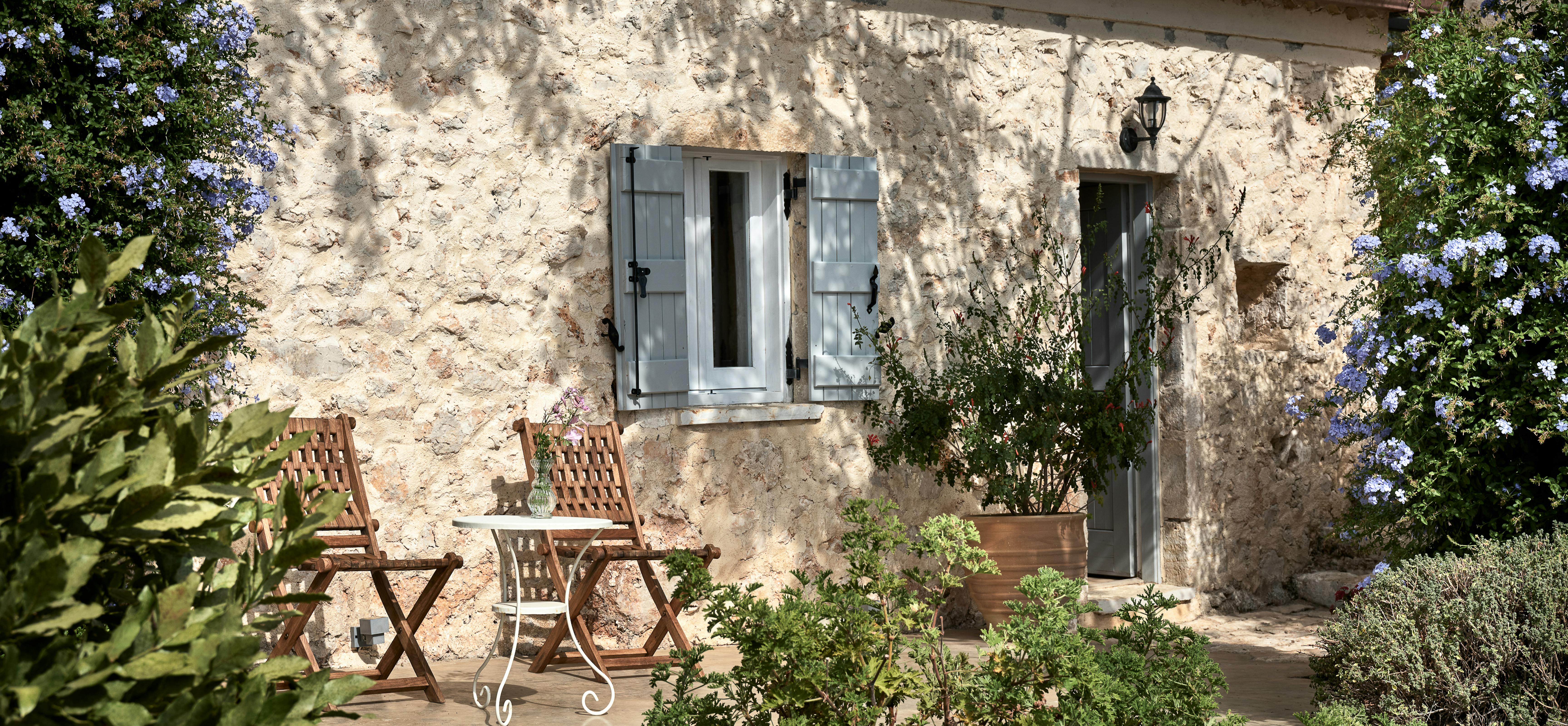 Weathered limestone farmhouse wall with traditional grey shutters framed by lush Mediterranean garden plantings. Simple wooden chairs suggest quiet contemplation spot among natural stone and greenery.
