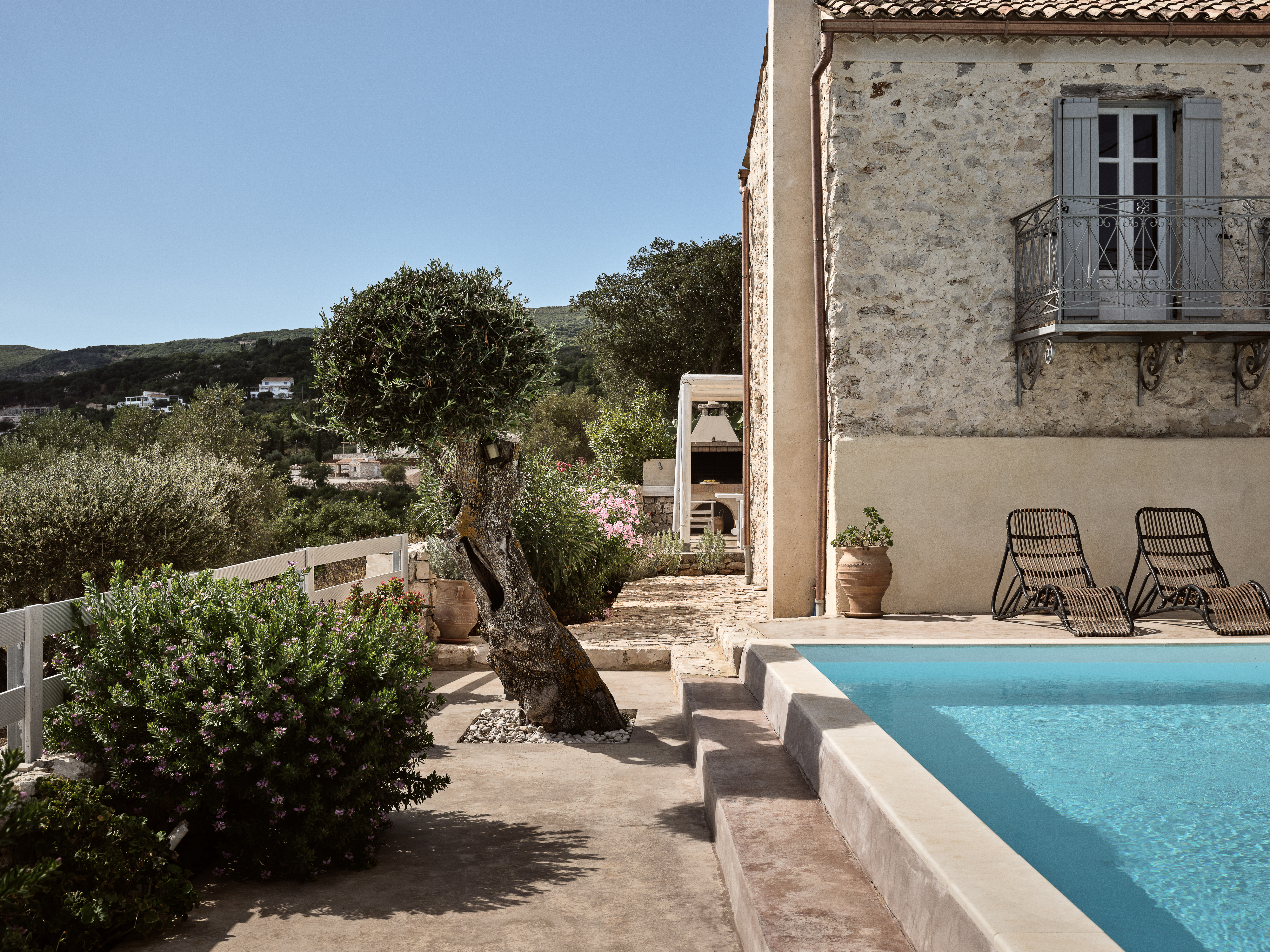 Traditional stone villa with wrought-iron balcony, traditional shutters, and swimming pool nestled in Mediterranean landscape. Ancient olive tree and flowering plants frame this authentic countryside retreat.