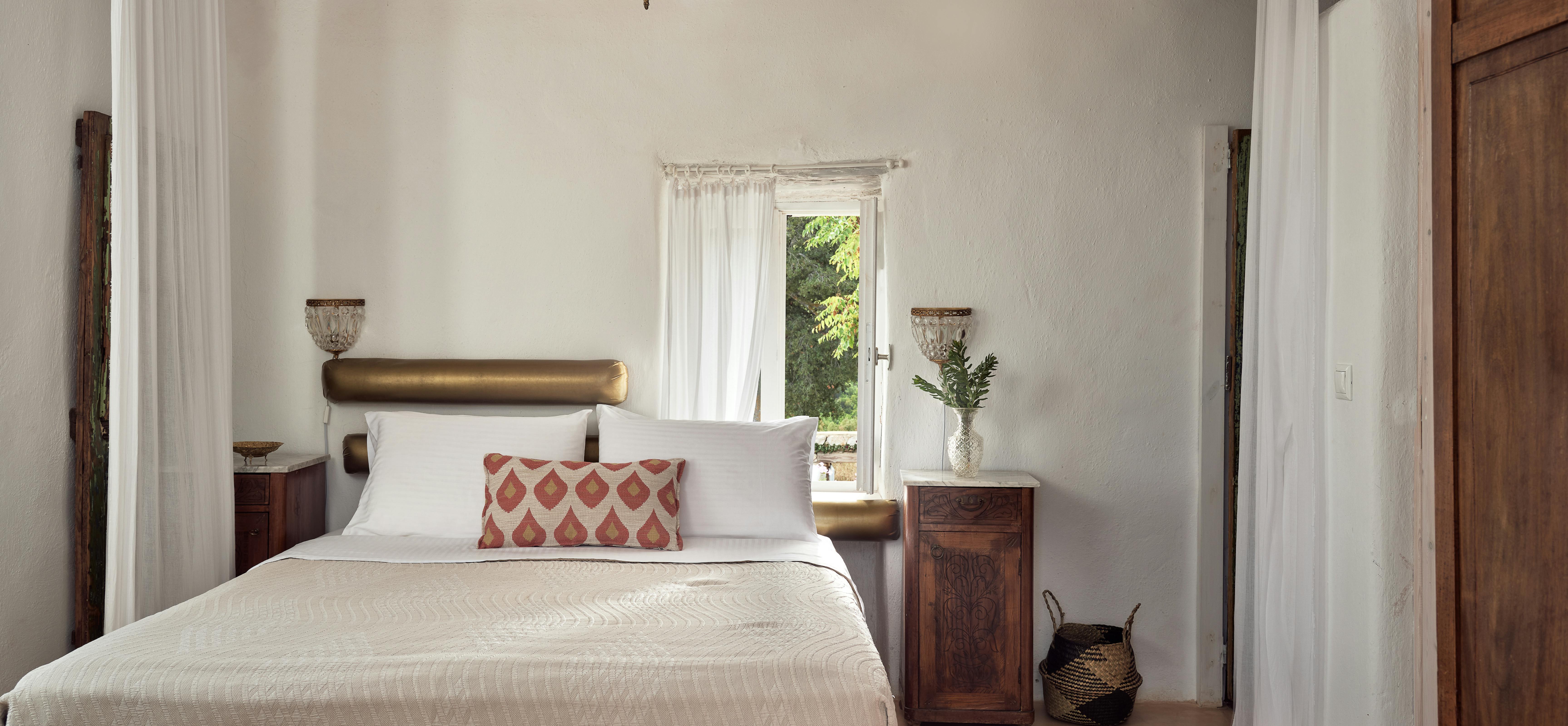 Romantic bedroom with brass headboard, crystal chandelier, and flowing white curtains under exposed wood ceiling. Antique nightstands and geometric pillow add sophisticated traditional touches.