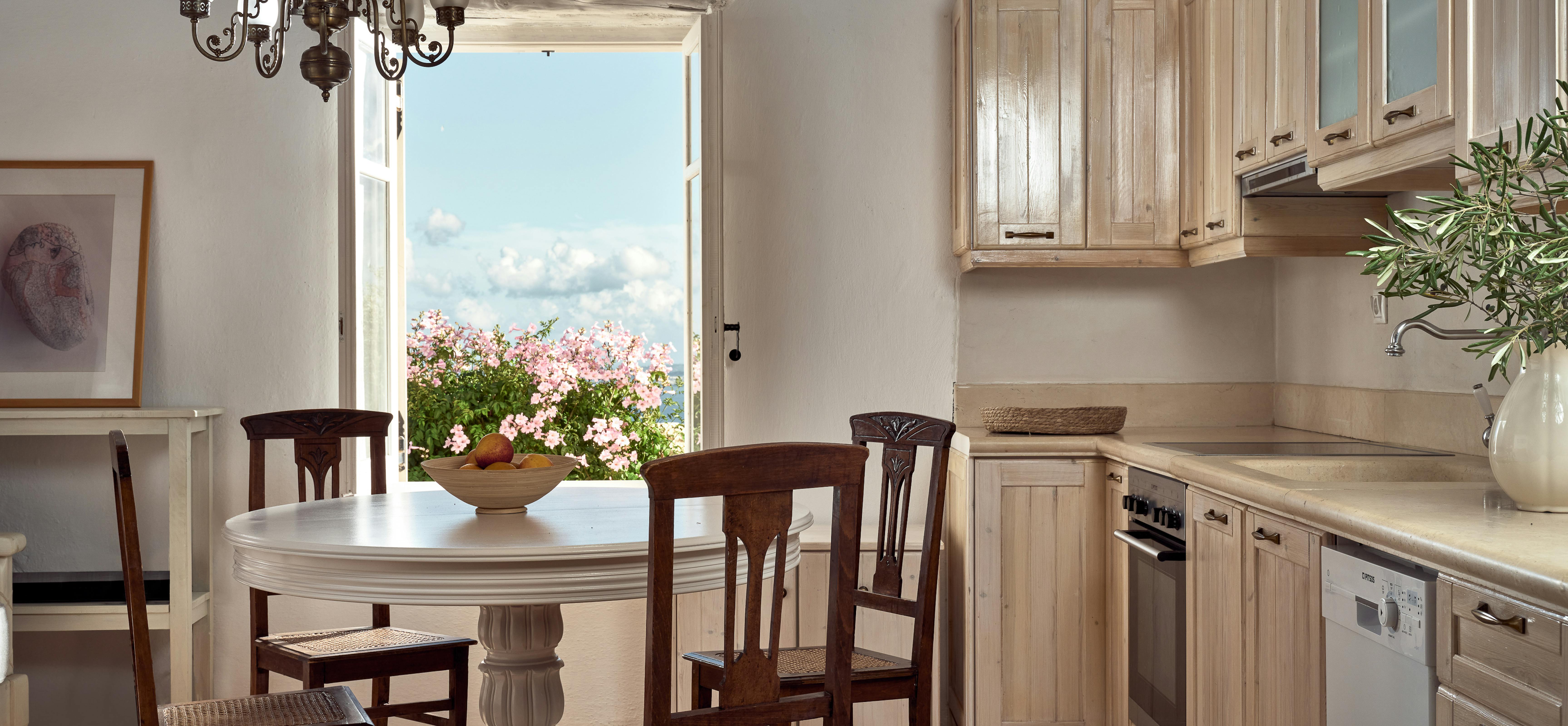 Traditional country kitchen with exposed beams, natural wood cabinetry, and wrought-iron chandelier overlooking flowering garden. Round white dining table with cane-seat chairs creates charming breakfast area.