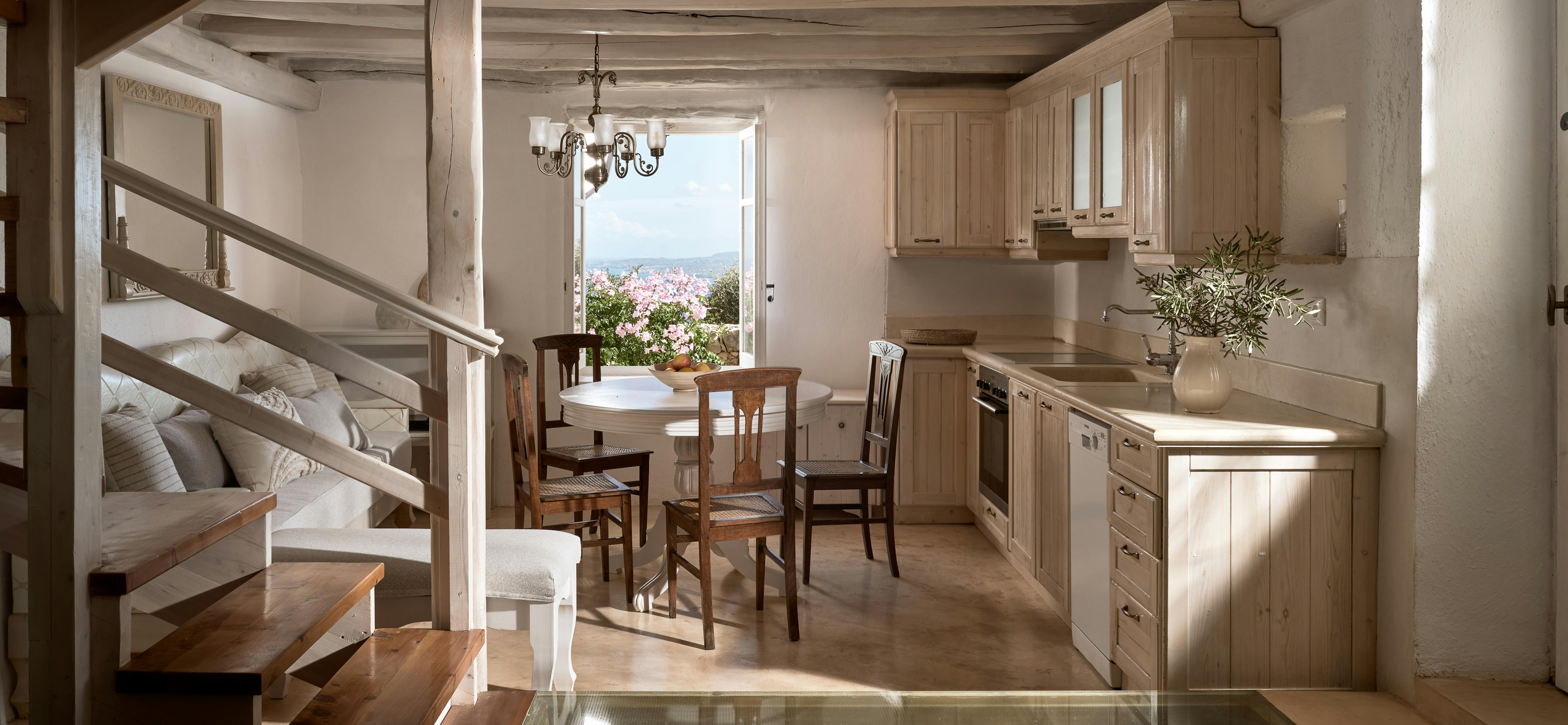 Rustic kitchen with exposed ceiling beams, natural wood cabinetry, and glass floor panel revealing ancient stone foundations. Wrought-iron chandelier illuminates dining area with garden views through French doors.