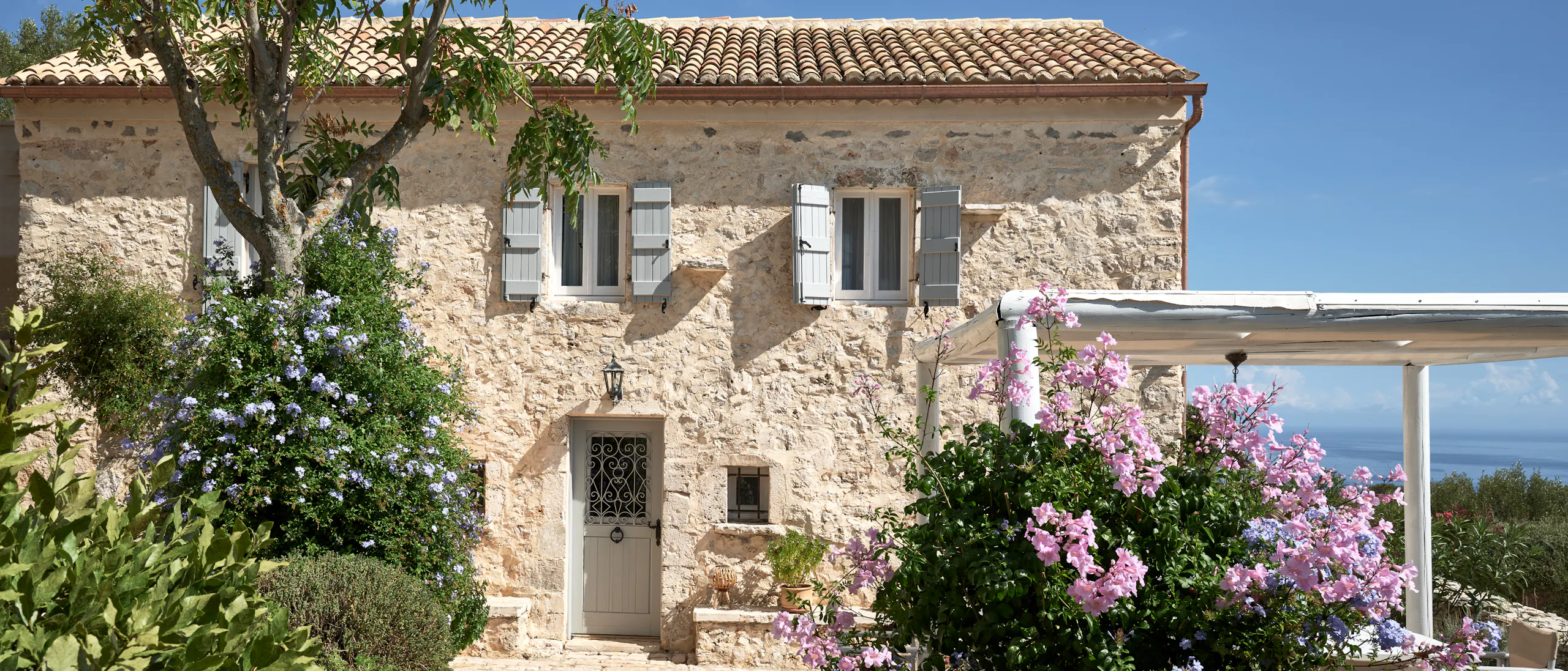 Traditional stone farmhouse with terracotta roof surrounded by vibrant purple and blue flowering plants. Cobblestone pathway leads to entrance with decorative ironwork door amid lush Mediterranean landscaping.