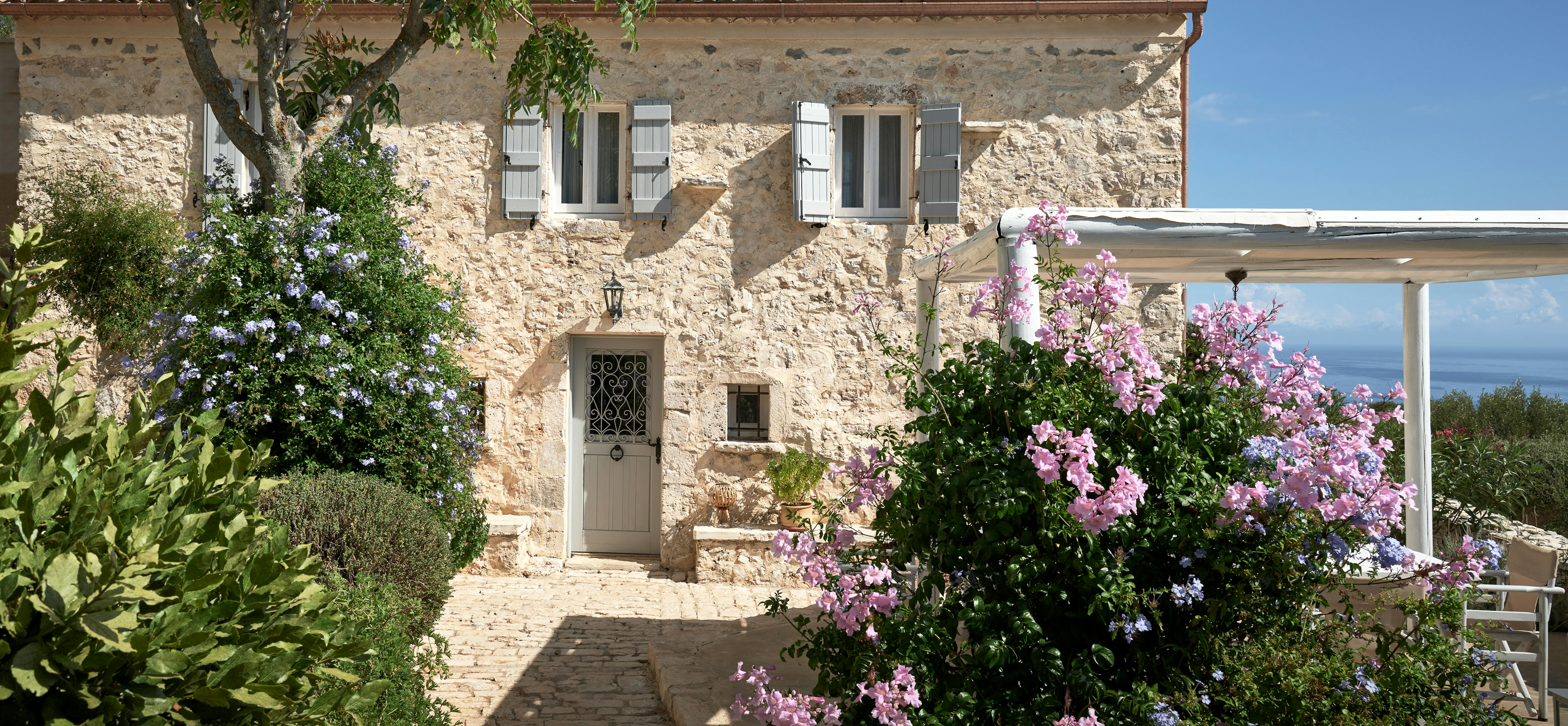 Traditional stone farmhouse with terracotta roof surrounded by vibrant purple and blue flowering plants. Cobblestone pathway leads to entrance with decorative ironwork door amid lush Mediterranean landscaping.
