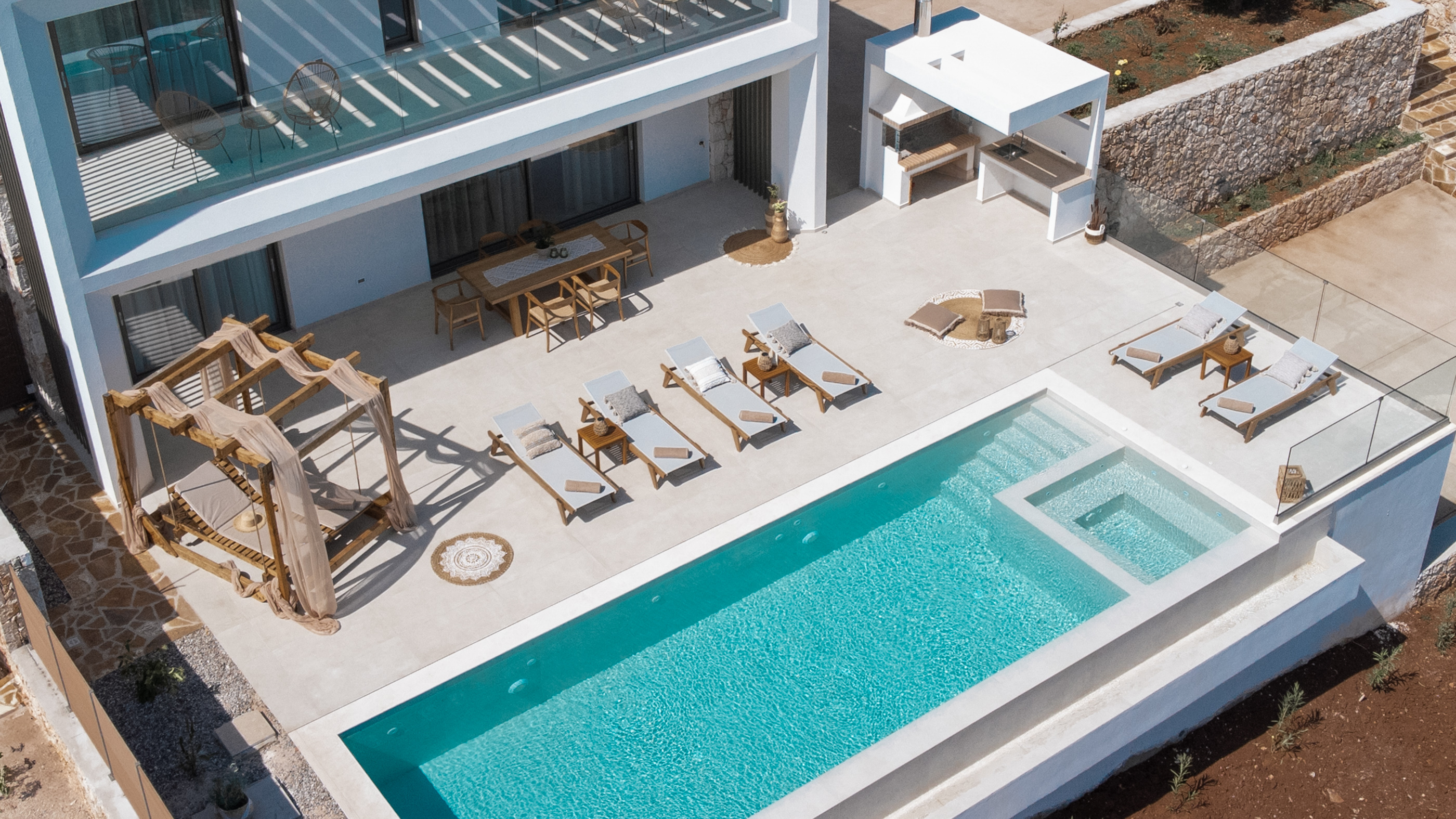 Aerial view of modern villa's pool deck with rectangular infinity pool, wooden sun loungers, and geometric outdoor furniture arrangement. Clean white architecture contrasts with surrounding landscape.