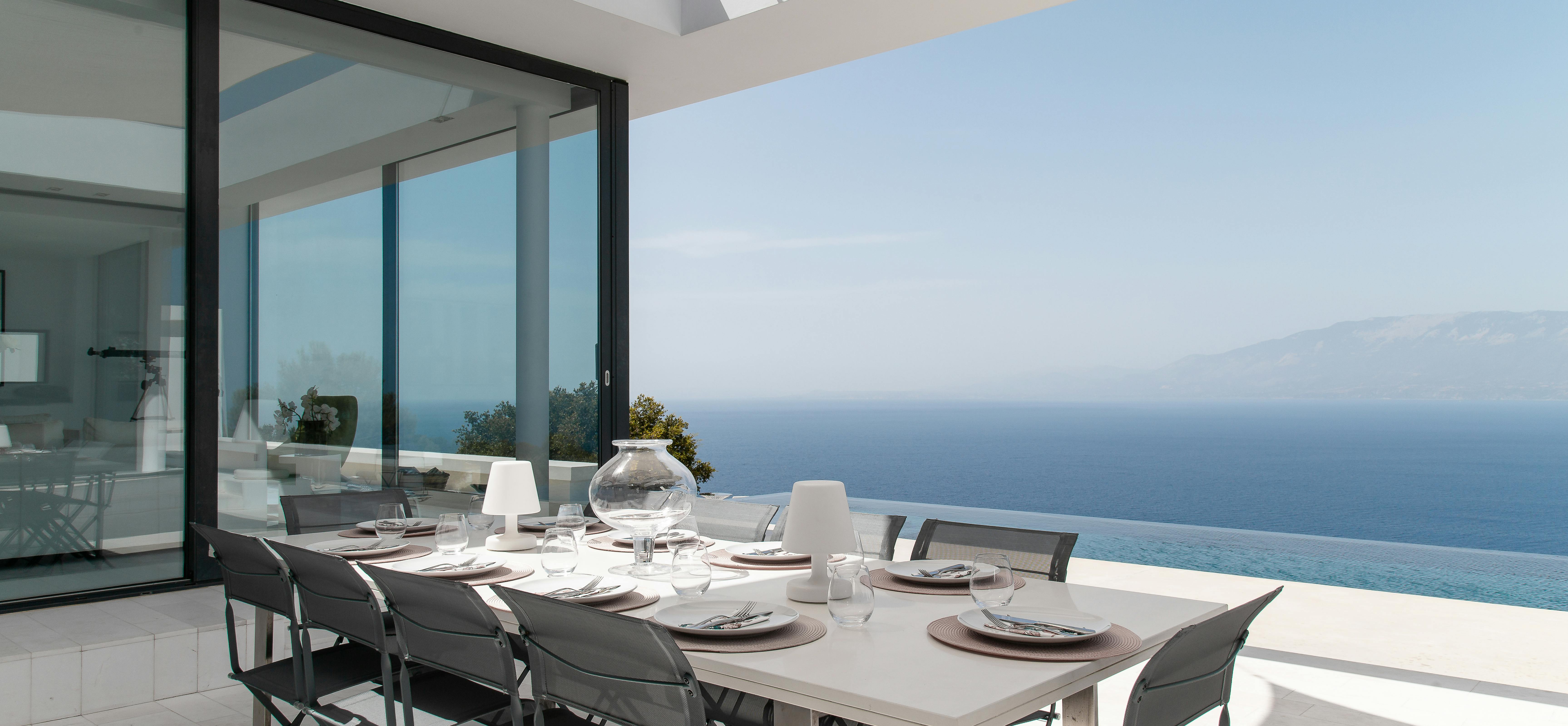 Covered terrace dining area with set table overlooking infinity pool and Mediterranean coastline. Perfect al fresco dining space with dramatic ocean backdrop.
