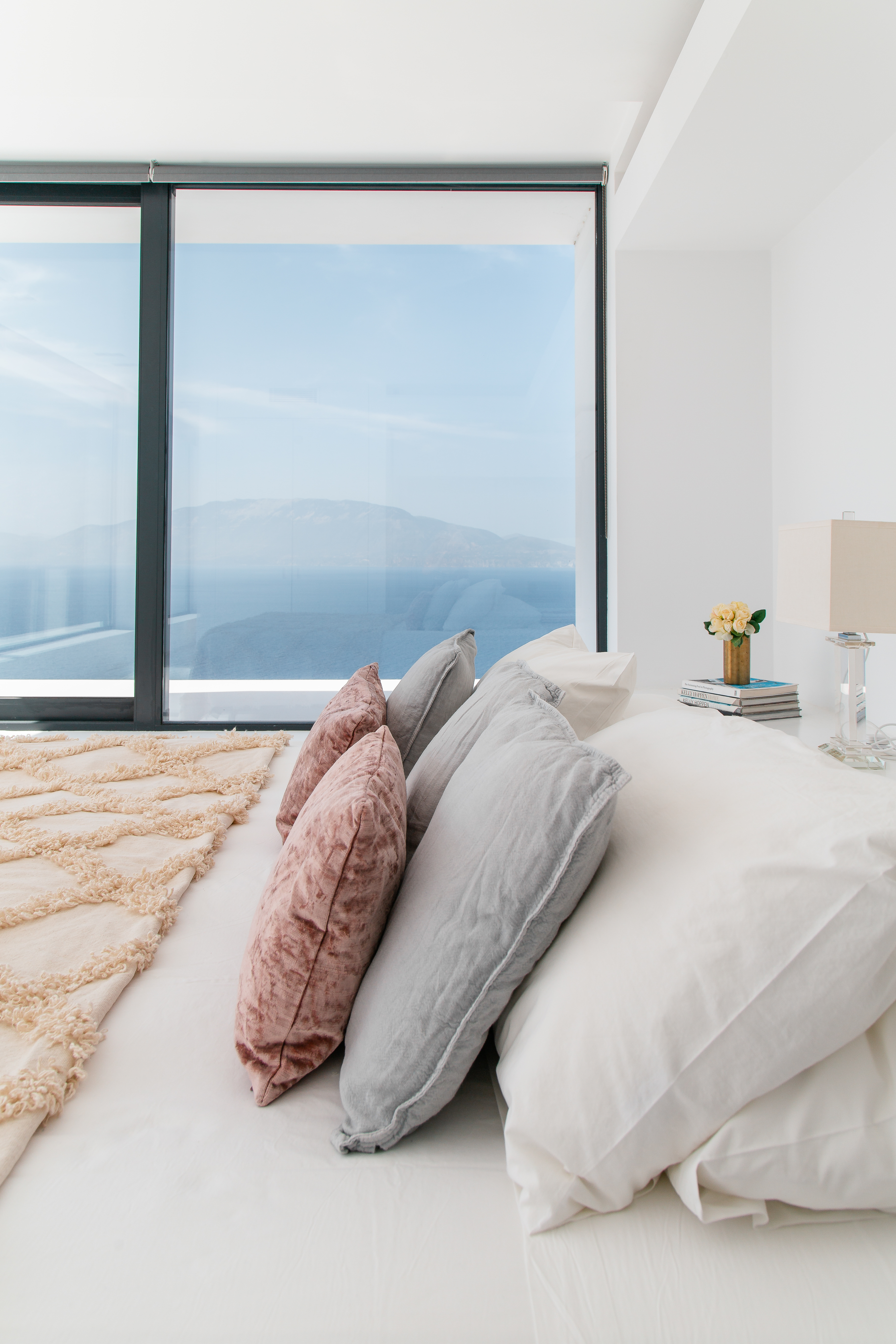 Luxurious bedroom with layered neutral bedding, pink accents, and expansive windows framing mountain and sea views. White interior emphasises natural beauty beyond
