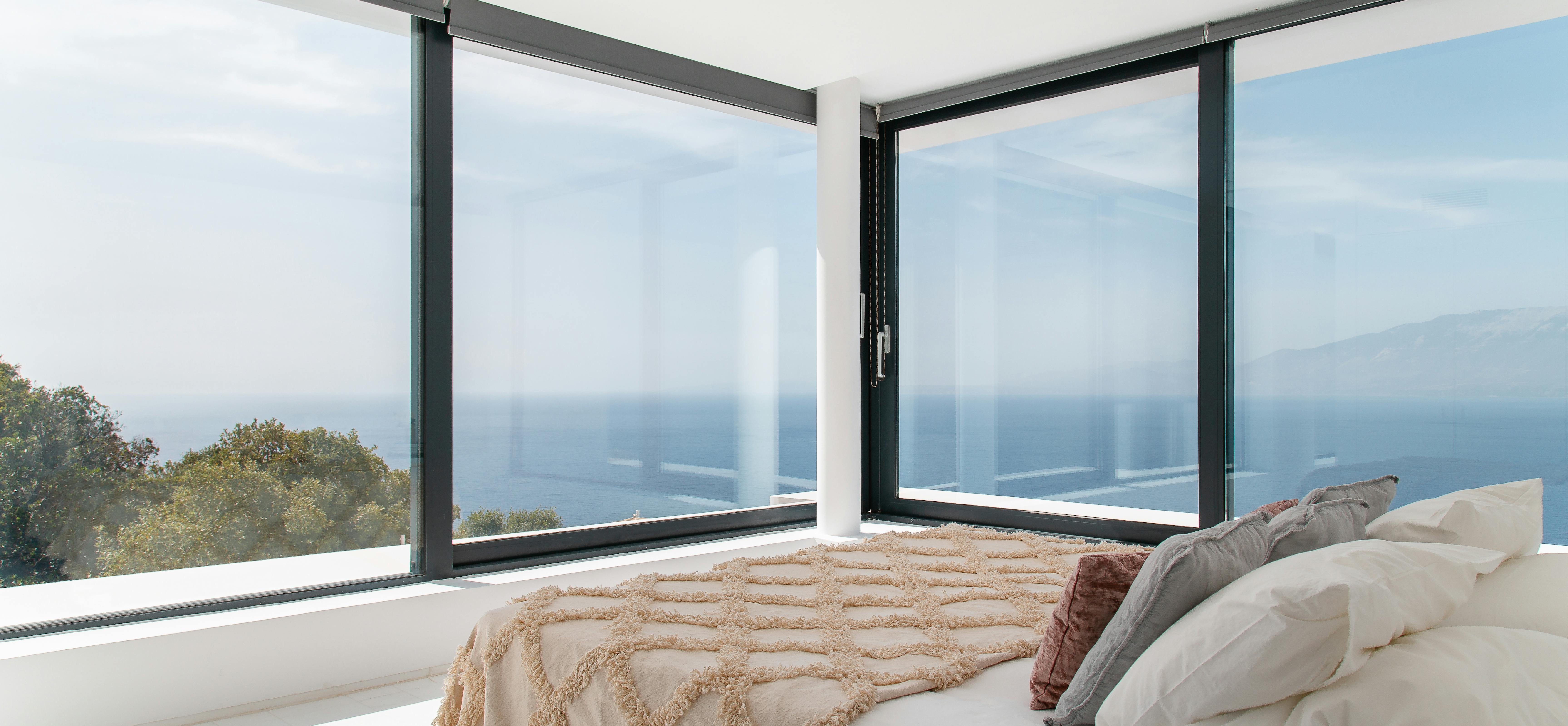 Master bedroom featuring panoramic windows, neutral bedding with geometric throw, and unobstructed ocean views. Minimalist design emphasises natural light and seascape vistas.
