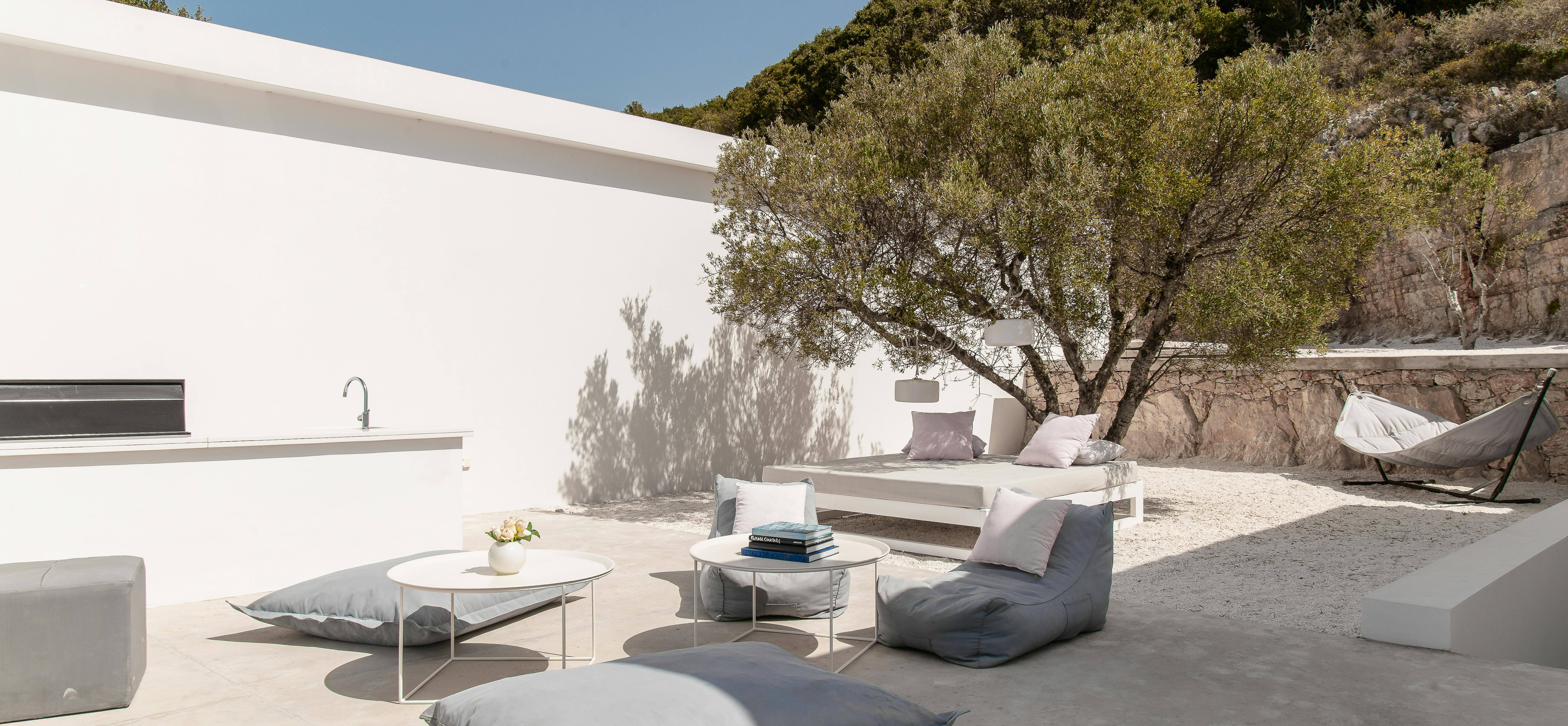 Outdoor lounge area with white furniture, grey floor cushions, and modern coffee table beneath natural tree canopy. Minimalist design blends seamlessly with hillside setting.