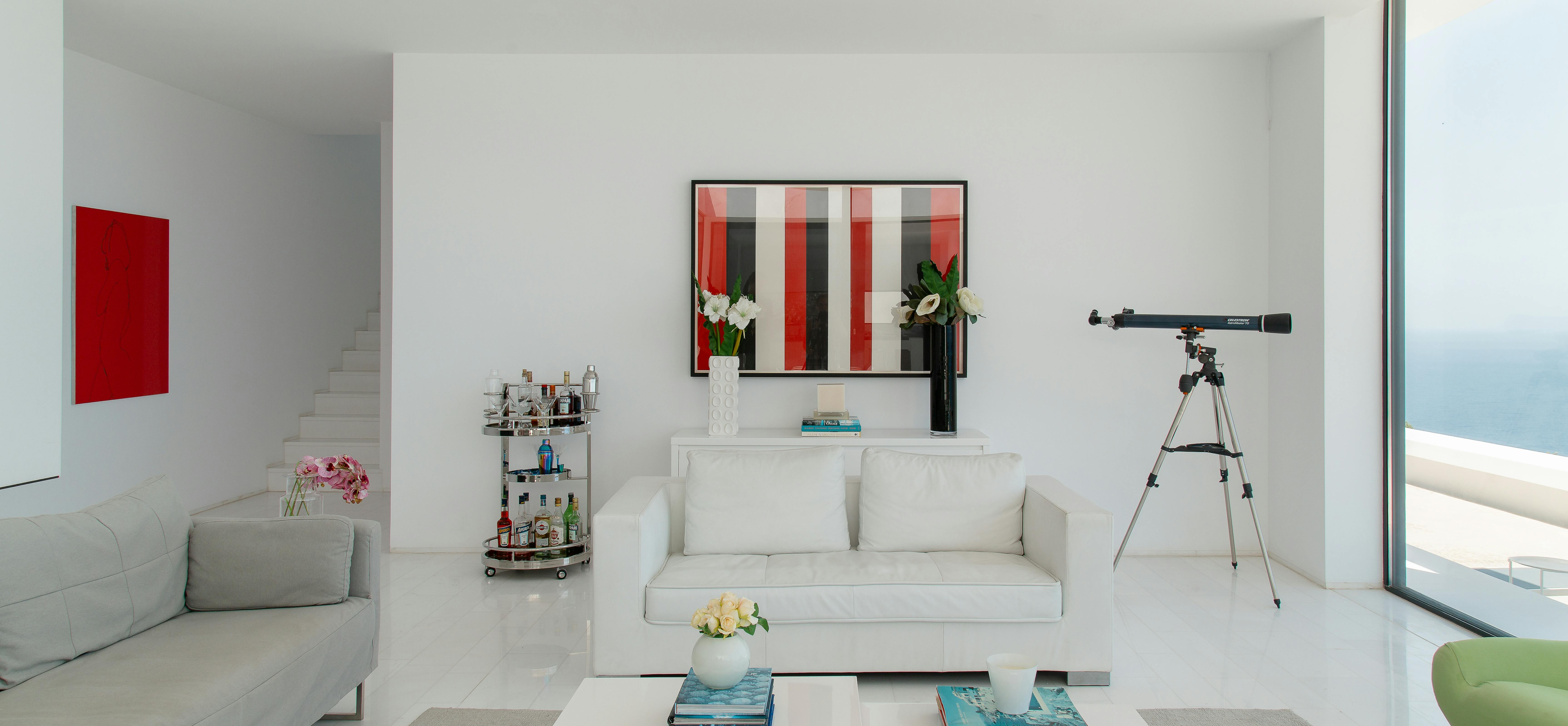 Minimalist living room with white sectional sofas, geometric coffee table, and telescope positioned near floor-to-ceiling windows. Red and striped artwork add colour to neutral palette.