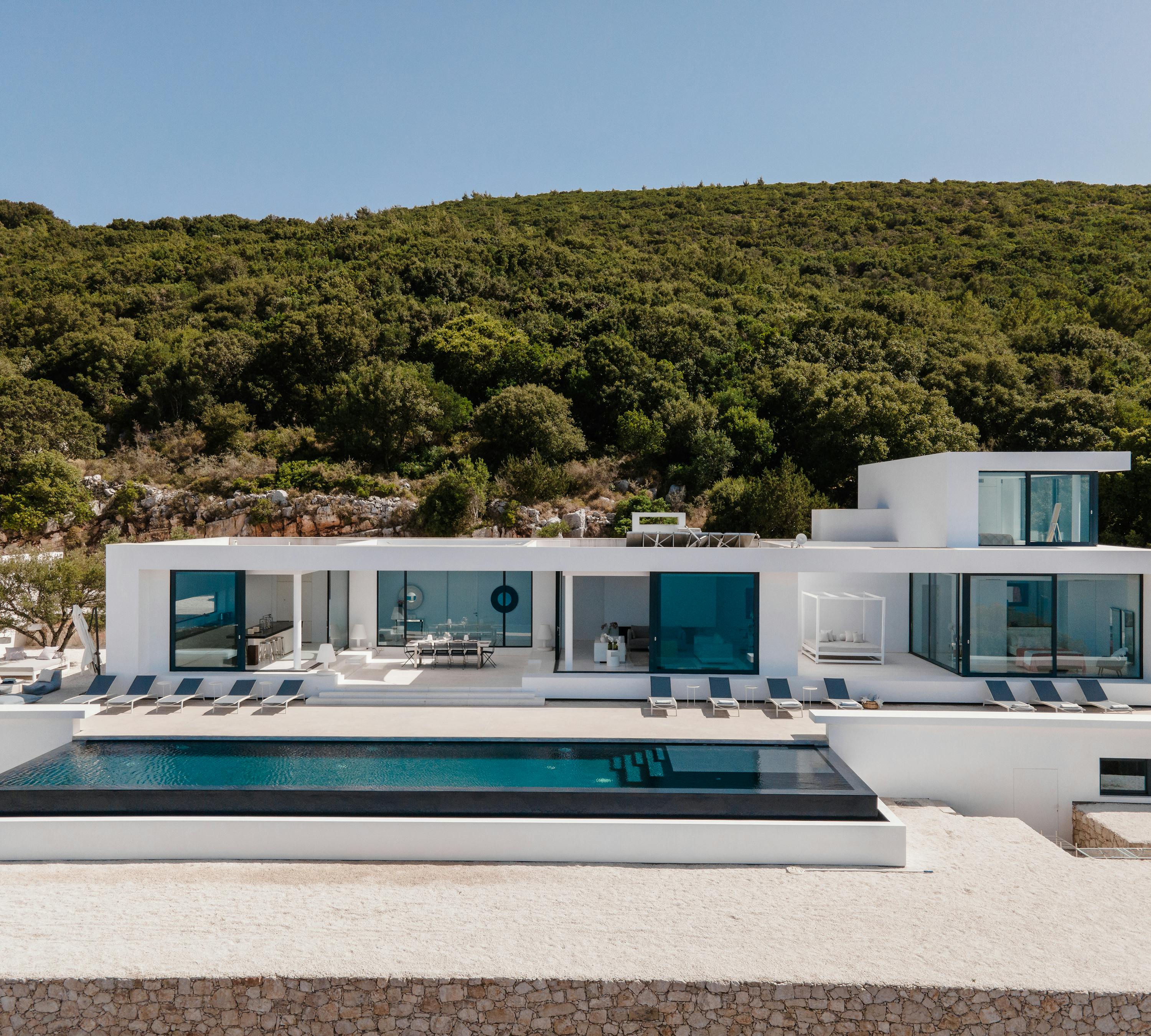 Modern white villa with geometric architecture featuring floor-to-ceiling glass walls and black-framed infinity pool set against forested hillside. Clean minimalist design emphasises indoor-outdoor living.