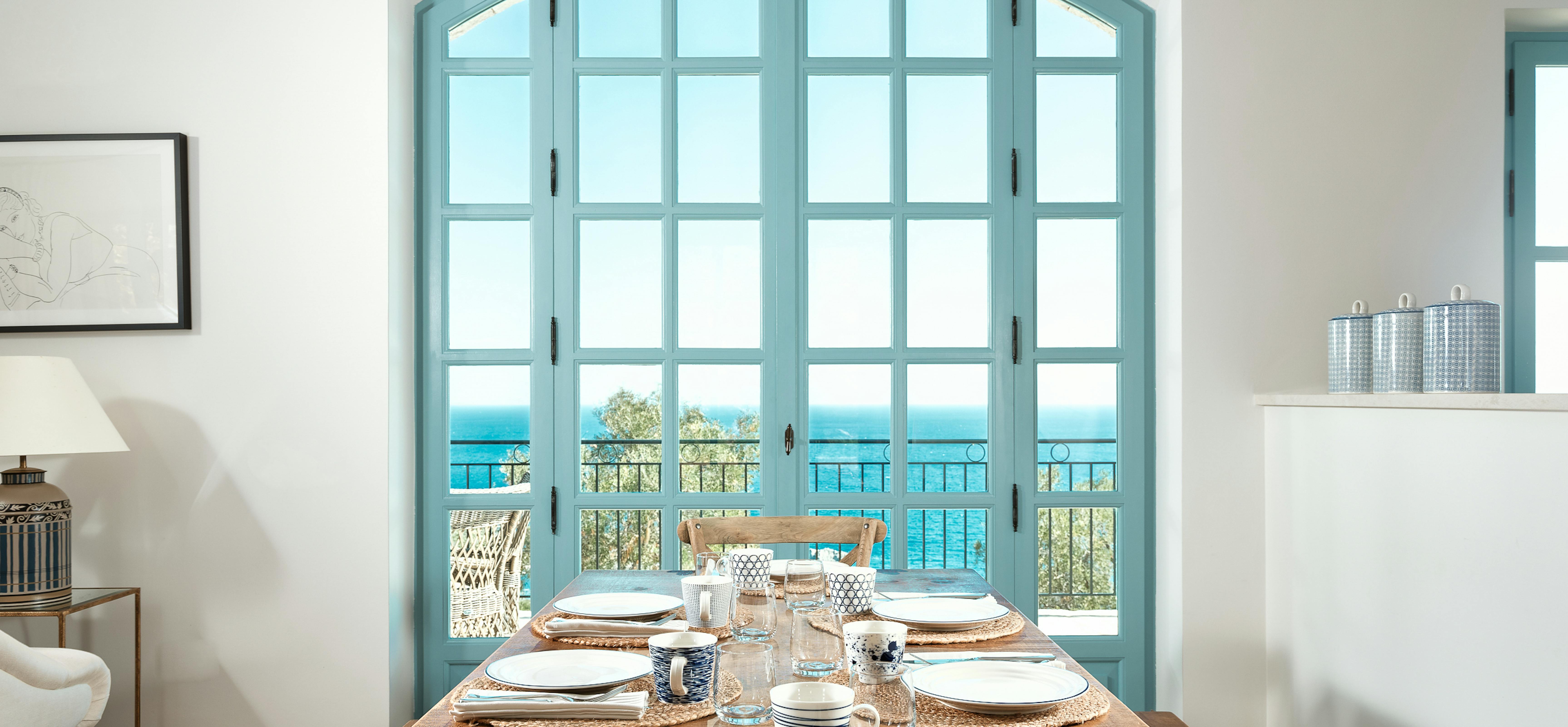 Mediterranean dining room with wooden table set for six, featuring turquoise French doors opening to ocean-view balcony. Woven placemats and white dinnerware create casual coastal elegance.