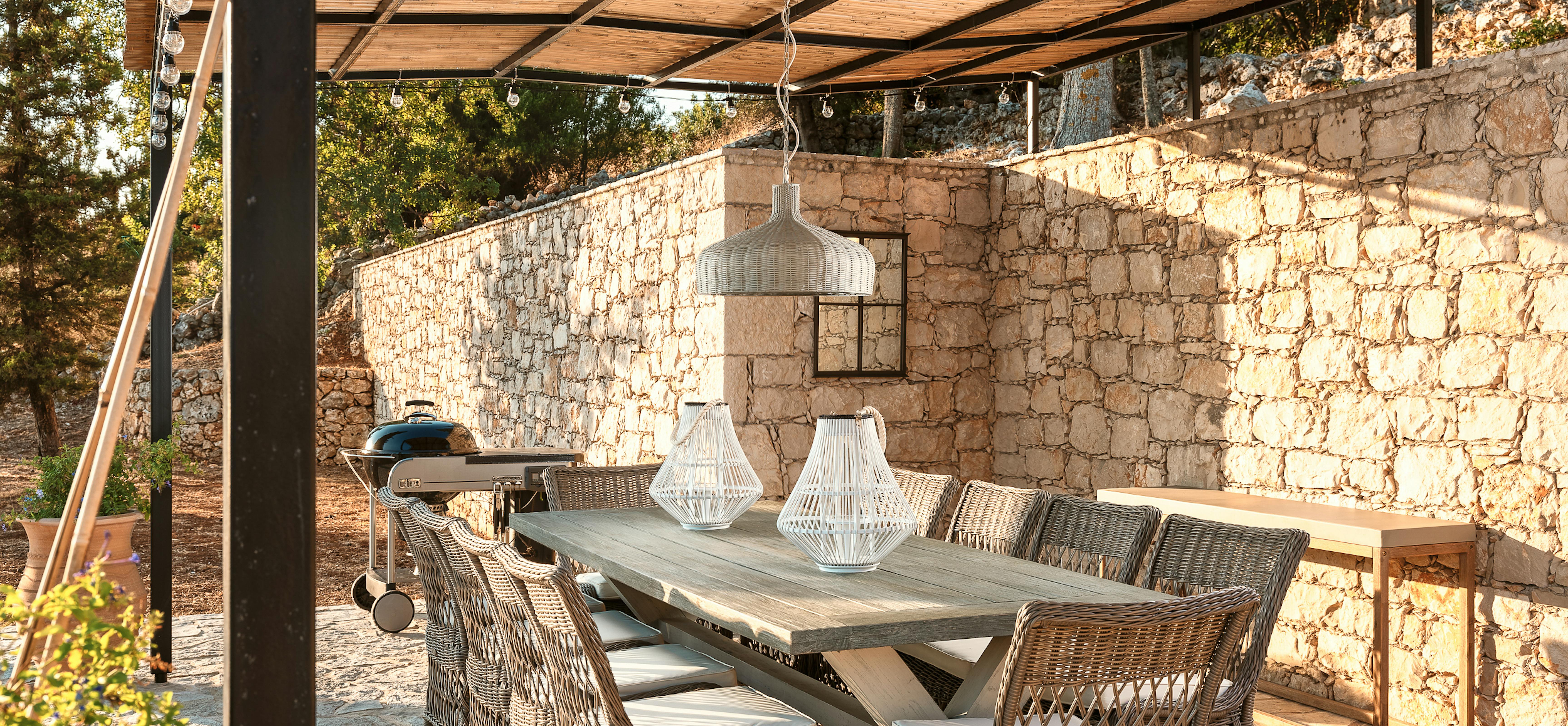 Covered outdoor dining area with woven furniture, rustic wooden table, and stone wall backdrop. String lights and natural materials create intimate evening atmosphere.
