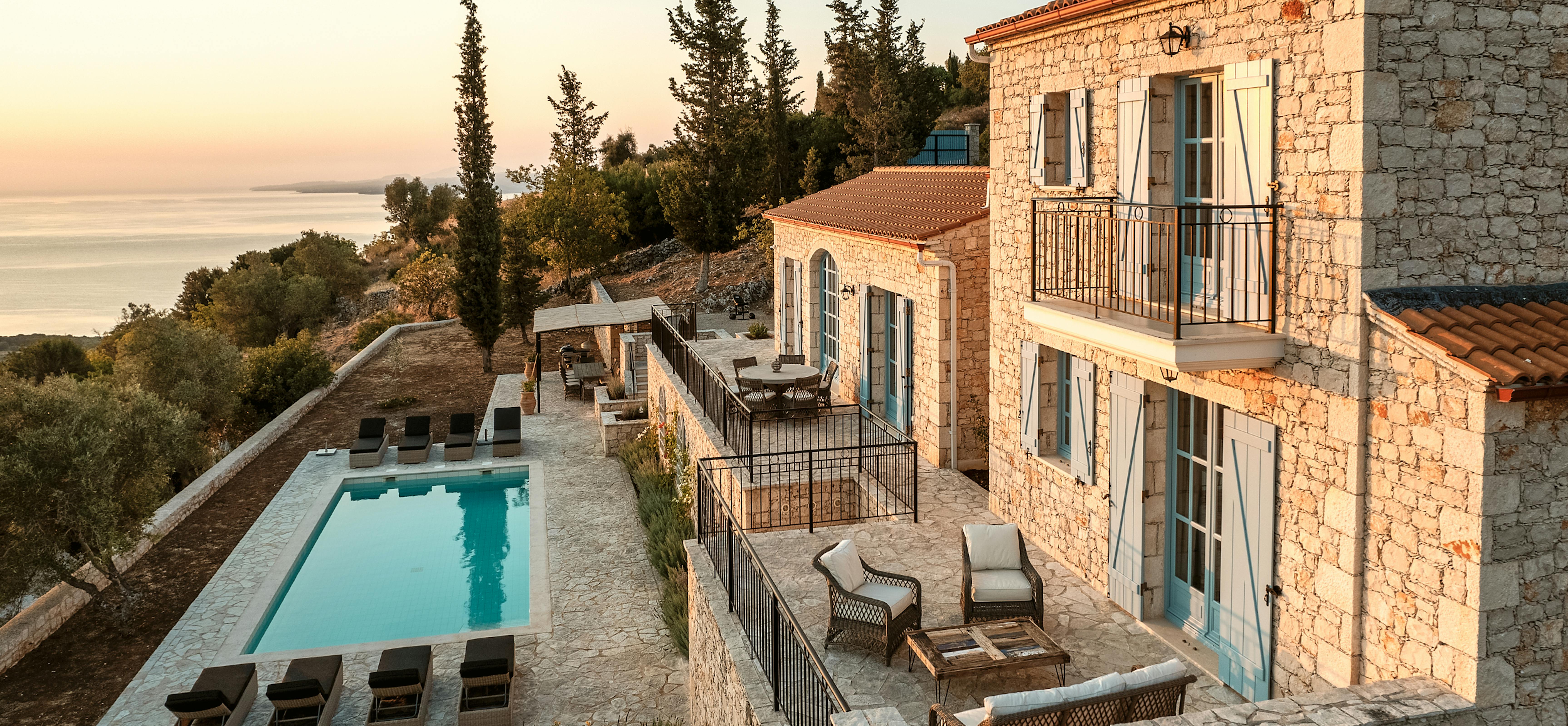 Traditional Mediterranean stone villa with terracotta roof overlooking sea at sunset, featuring multiple terraces, infinity pool, and turquoise shutters. Warm golden light illuminates the limestone architecture and cypress trees.