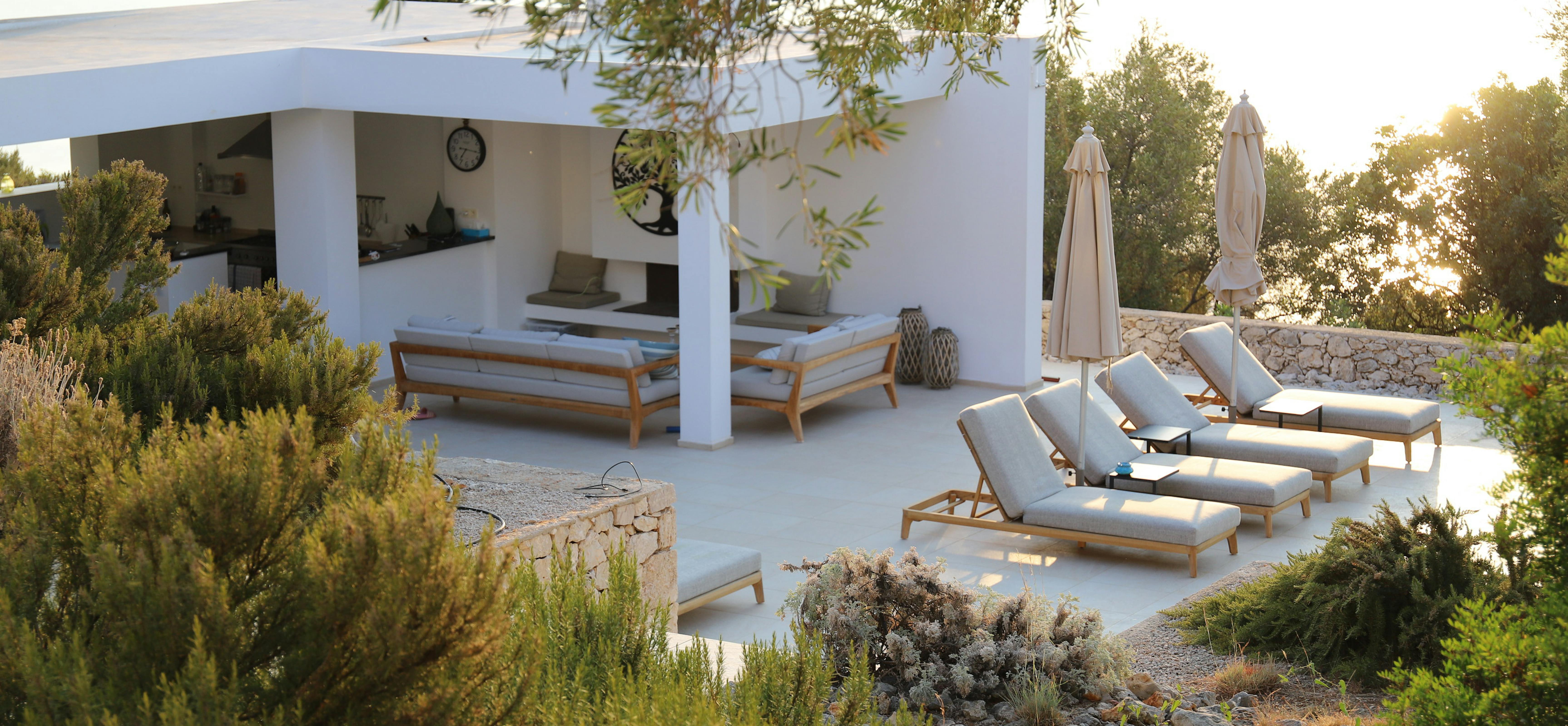 Covered outdoor lounge area with wooden furniture, umbrellas, and Mediterranean landscape at sunset.