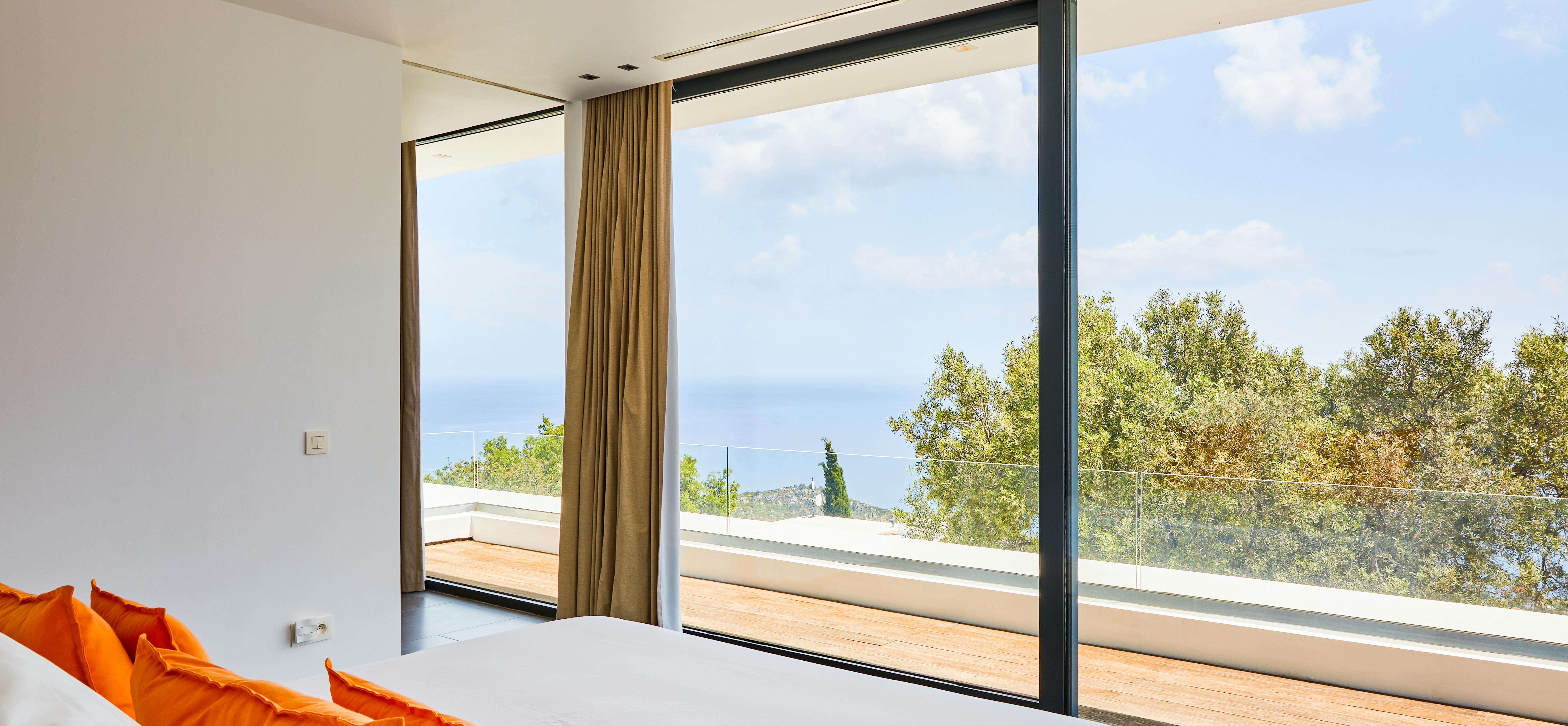 Minimalist bedroom with orange accent pillows, large windows, and view of wooden terrace outside.