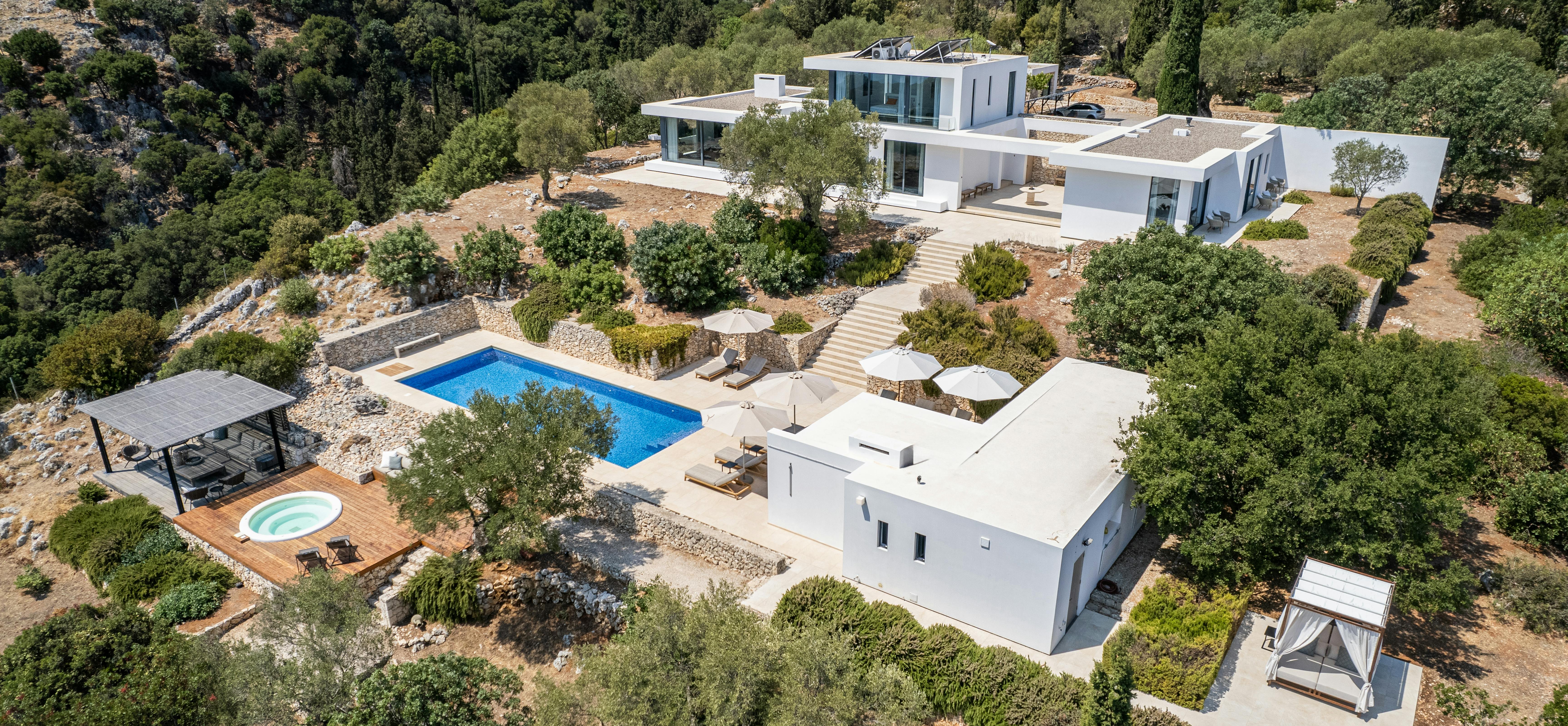 Aerial view of contemporary white villa complex with pools and terraced landscaping in forested hills.