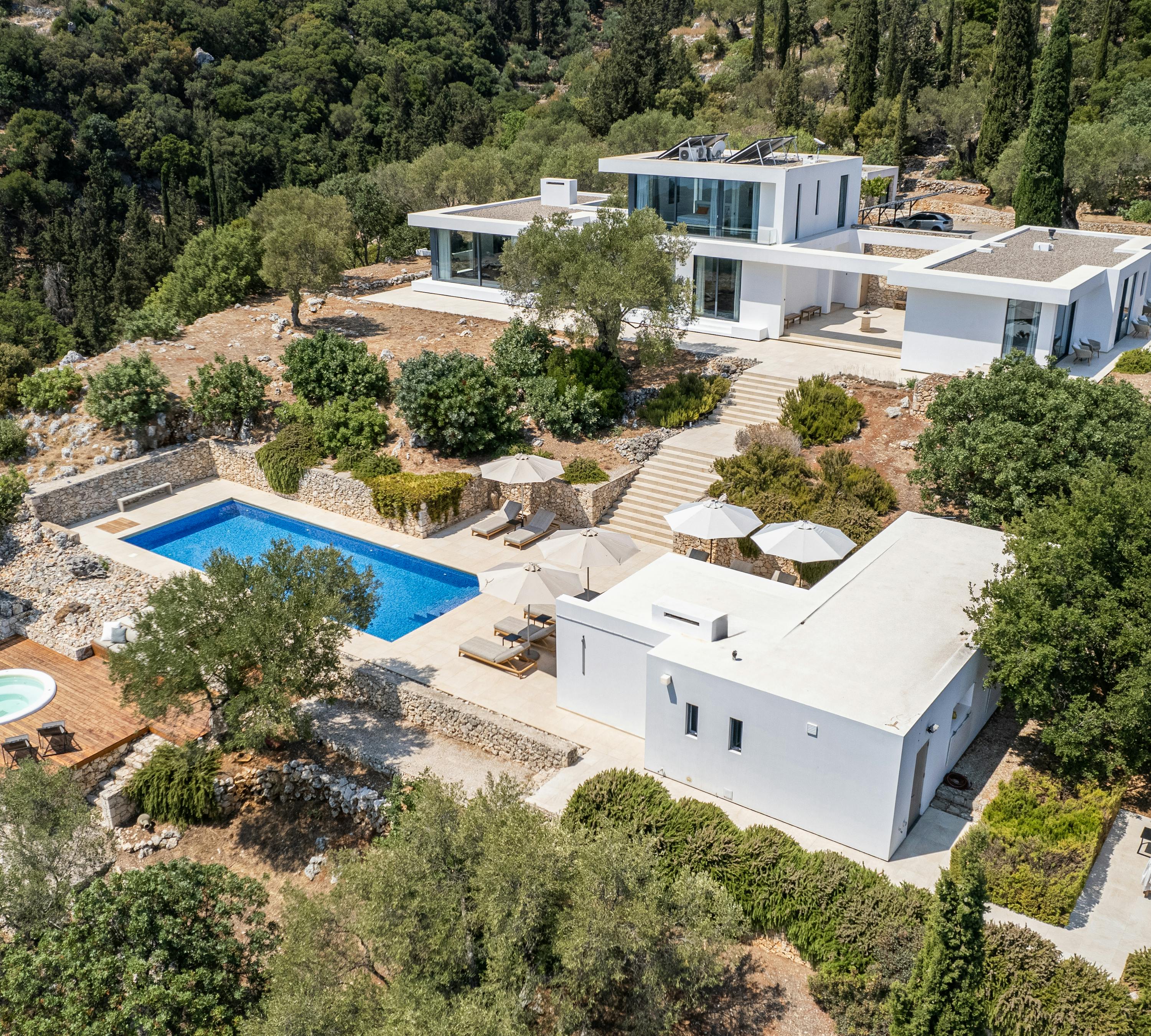Aerial view of contemporary white villa complex with pools and terraced landscaping in forested hills.