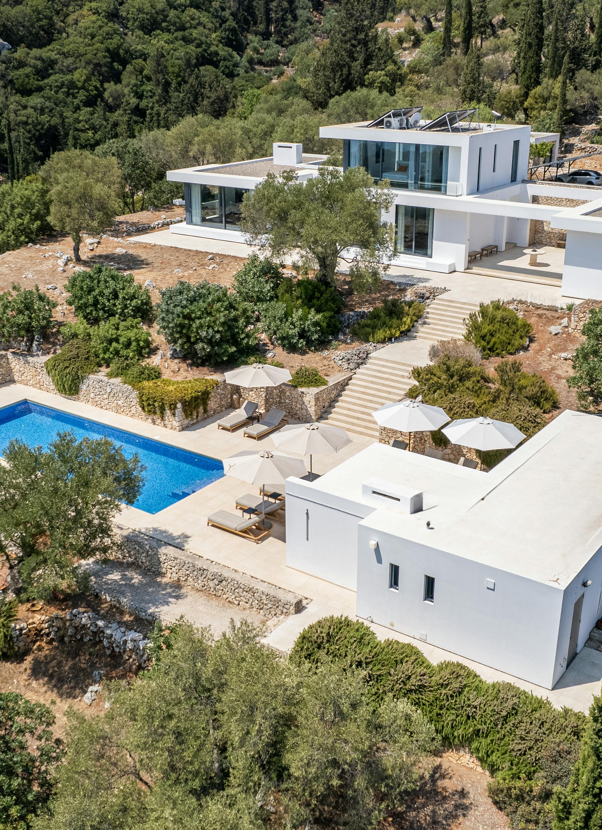 Aerial view of contemporary white villa complex with pools and terraced landscaping in forested hills.