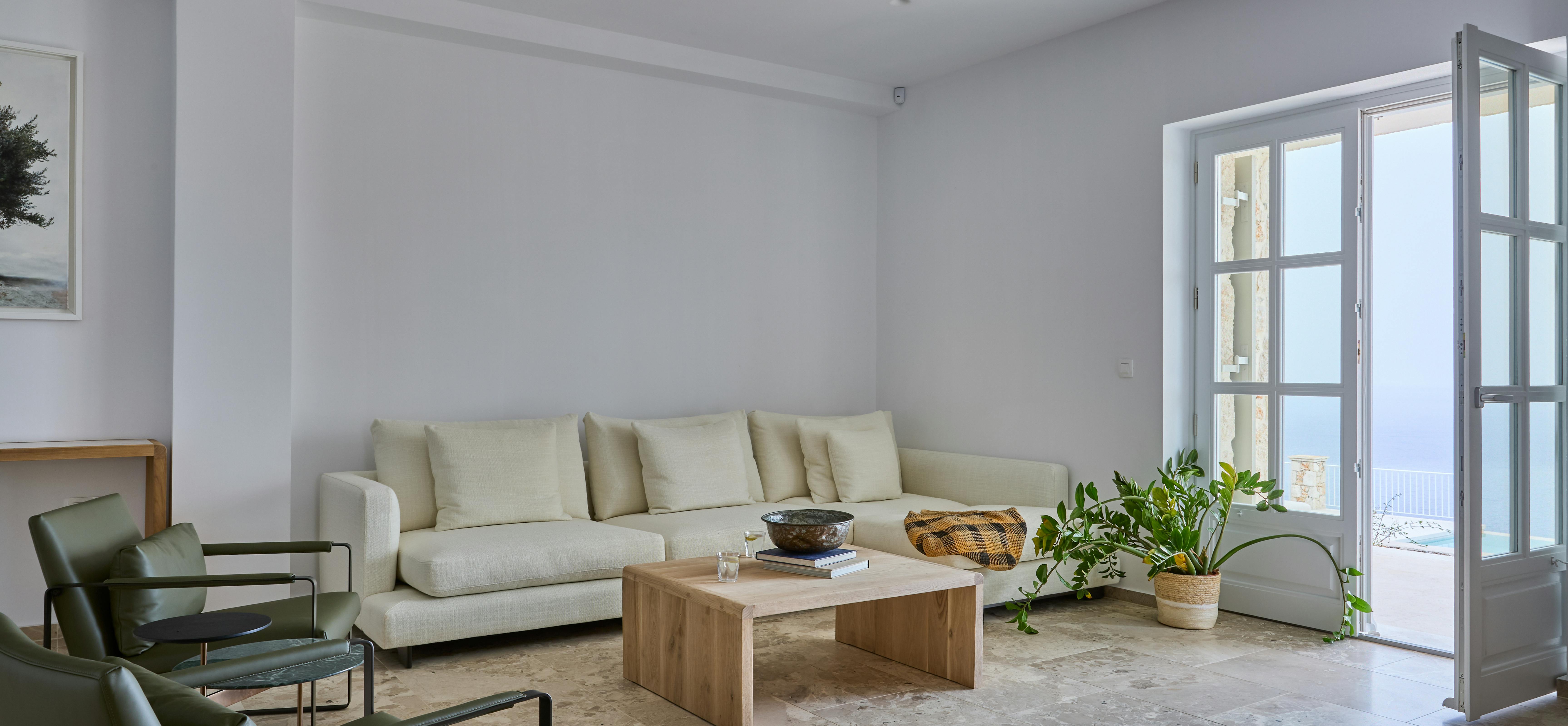 The main living area features neutral-toned sectional seating and a timber coffee table on polished stone floors. Large French doors connect the indoor space to outdoor terraces with Mediterranean views.