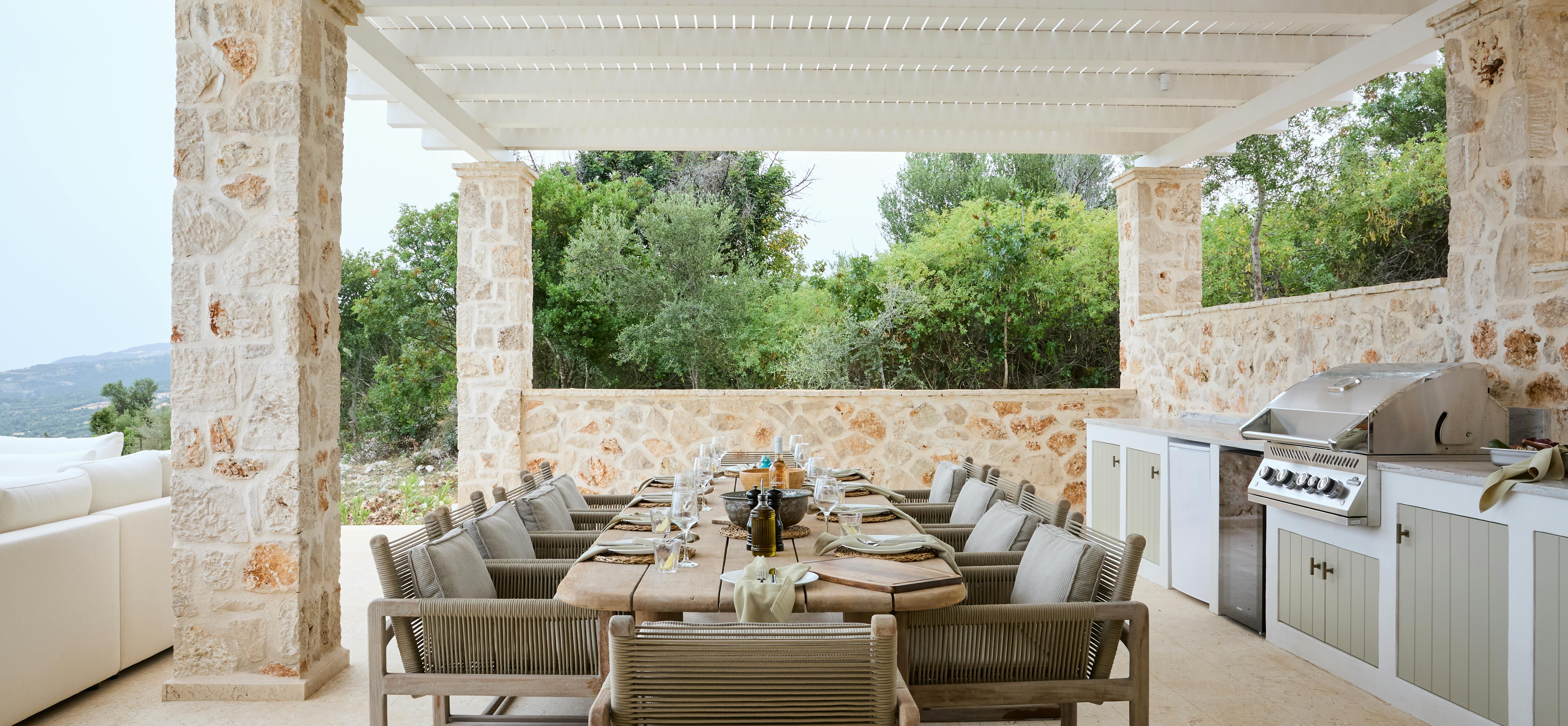 An outdoor covered dining area features a large table set for entertaining under white timber pergola beams. The space includes built-in stone barbecue facilities and overlooks the Mediterranean landscape.
