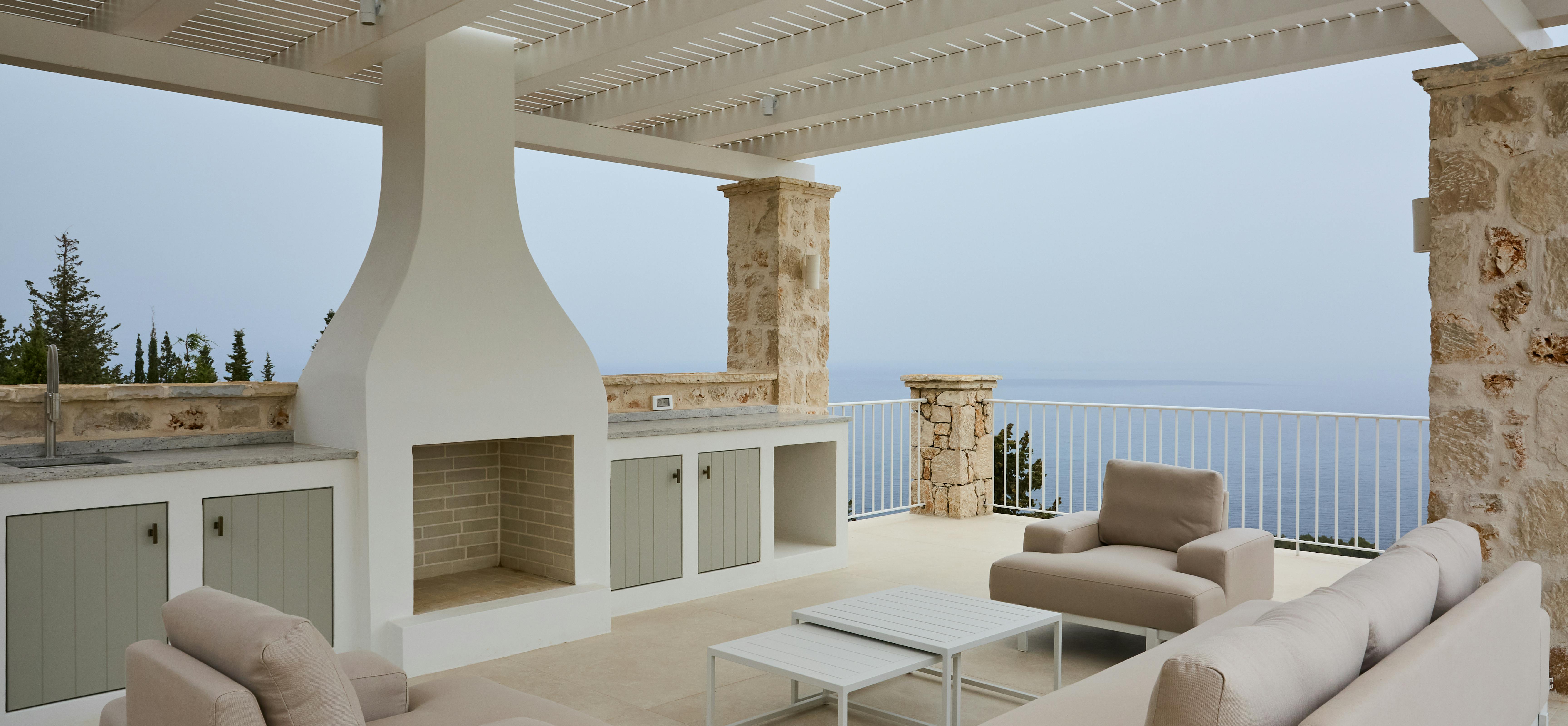 A sophisticated outdoor lounge area beneath a white timber pergola includes built-in kitchen facilities and comfortable seating. The space combines cooking, dining and relaxation areas with sweeping sea views as the backdrop.