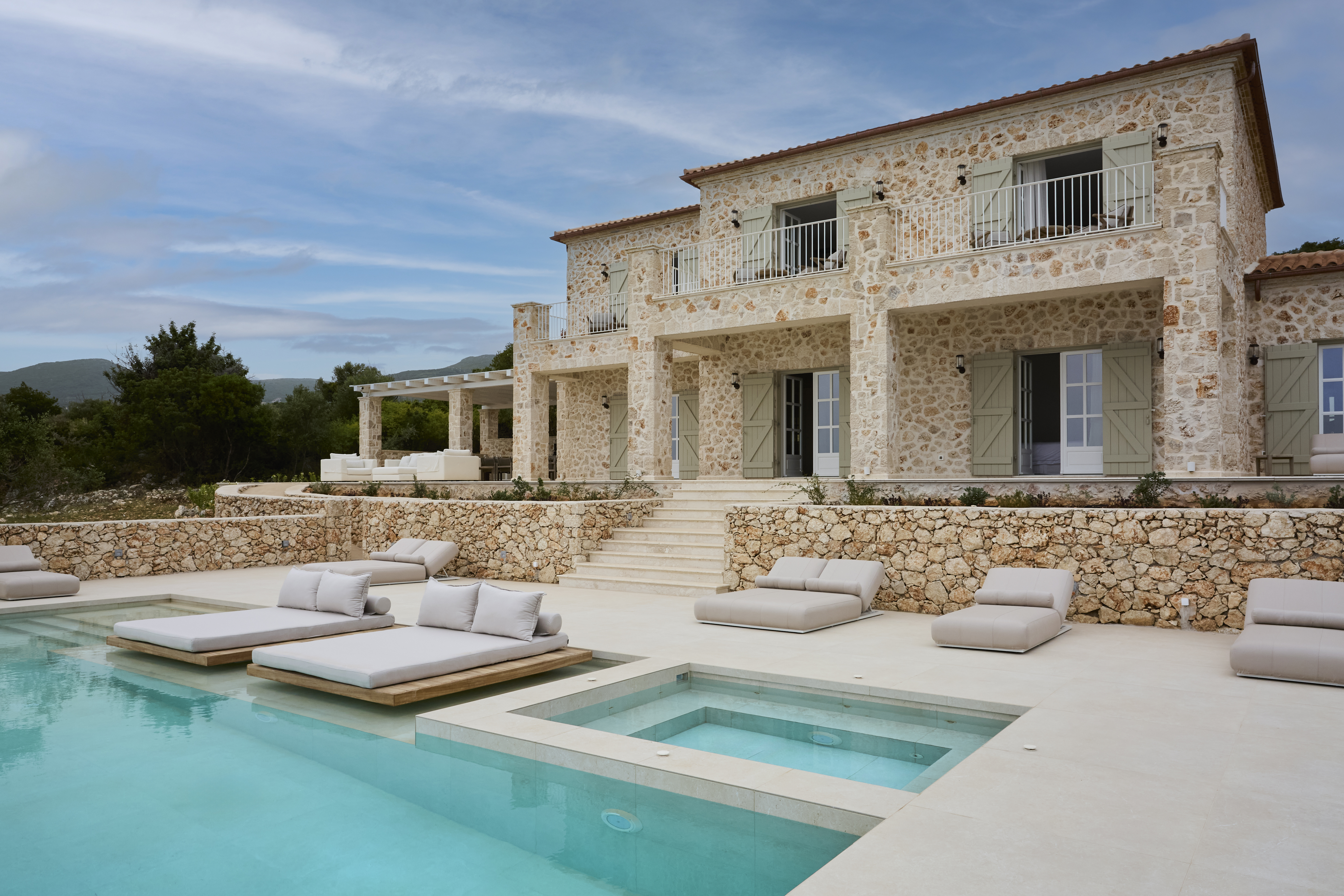 The villa's impressive stone facade showcases traditional Mediterranean architecture with sage green shutters and terracotta roof tiles. Multiple levels of terracing lead down to the infinity pool area with its luxury outdoor furniture.