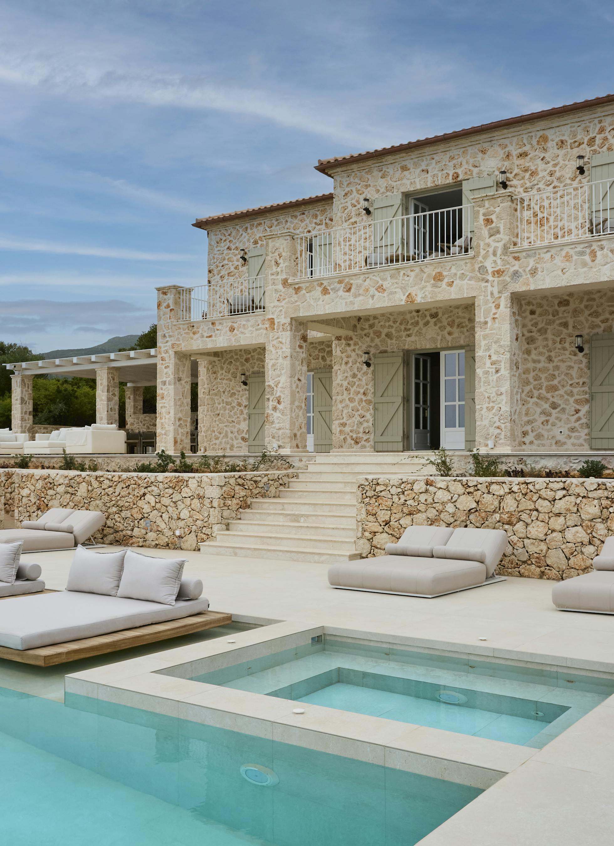 The villa's impressive stone facade showcases traditional Mediterranean architecture with sage green shutters and terracotta roof tiles. Multiple levels of terracing lead down to the infinity pool area with its luxury outdoor furniture.
